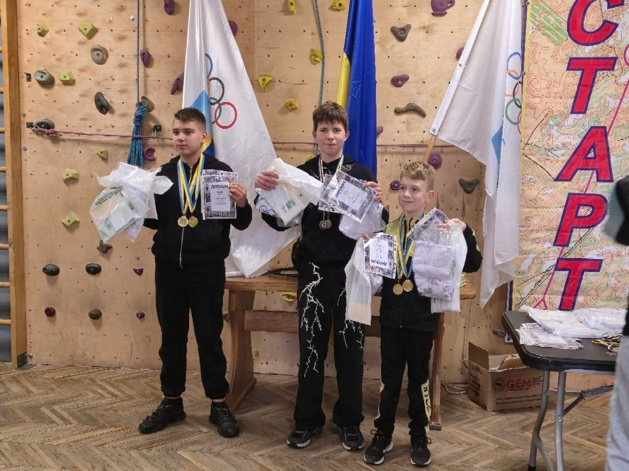 На світлині показано трьох юних призерів мінітурніру зі спортивного туризму на Херсонщині, які урочисто позують під час церемонії нагородження. Хлопці вишикувалися в один ряд на тлі дерев'яного скеледрому з різнокольоровими зачепами, тримаючи в руках завойовані дипломи, білі пакунки з призами та фірмовими футболками. Ліворуч стоїть підліток у чорному спортивному костюмі з двома золотими медалями на жовто-блакитних стрічках; по центру перебуває хлопець у чорній кофті та штанях із білим принтом блискавок, на шиї якого виблискує срібна медаль; а праворуч радісно посміхається наймолодший учасник у чорній олімпійці, який також виборов дві золоті нагороди. За спинами юних спортсменів встановлено великий Державний Прапор України та два білі прапори з олімпійськими кільцями, а праворуч височіє вертикальний банер із яскравим написом «СТАРТ» та топографічною картою місцевості.