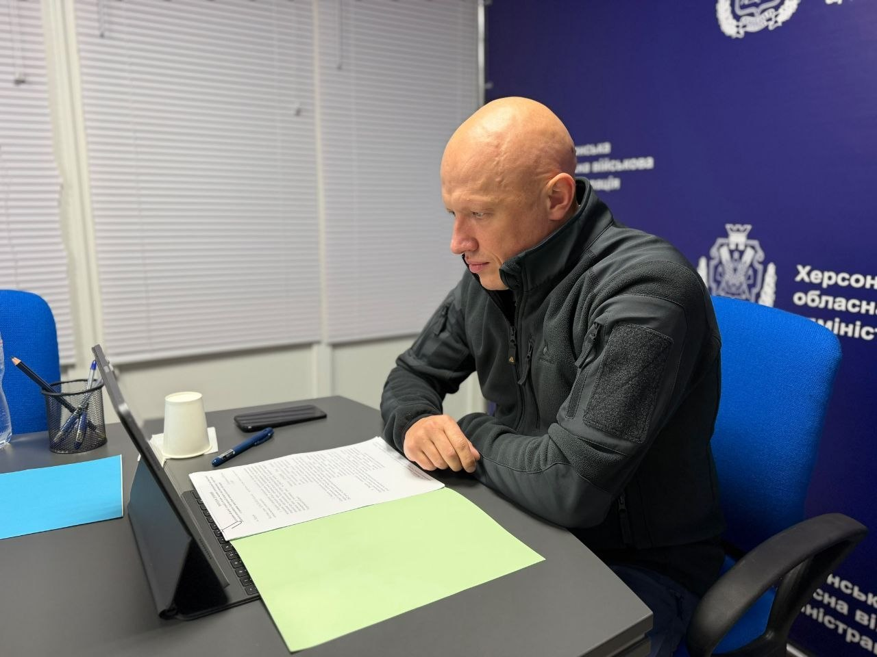 На світлині показано робочий момент засідання обласної комісії під головуванням заступника начальника Херсонської ОВА Євгенія Ігнатенка. У кабінеті за сірим офіційним столом сидить заступник голови ОВА — чоловік, одягнений у тактичну темно-сіру флісову кофту на блискавці із велкро-панелями (липучками) на рукавах. Він зосереджено схилився над розгорнутими на світло-зеленій папці паперовими документами зі зверненнями громадян, тримаючи руки на столі перед відкритим чорним планшетом-ноутбуком на підставці. На робочій поверхні перед ним також лежать синя кулькова ручка, чорний смартфон, стоїть білий паперовий стаканчик та сітчастий стакан-органайзер із олівцями. Зустріч проходить у приміщенні адміністрації на тлі синьої стіни з емблемами та білими офіційними написами, де чітко видно частину тексту «Херсонська обласна військова адміністрація», а ліворуч у кімнаті облаштовано великі вікна із закритими білими горизонтальними жалюзі.