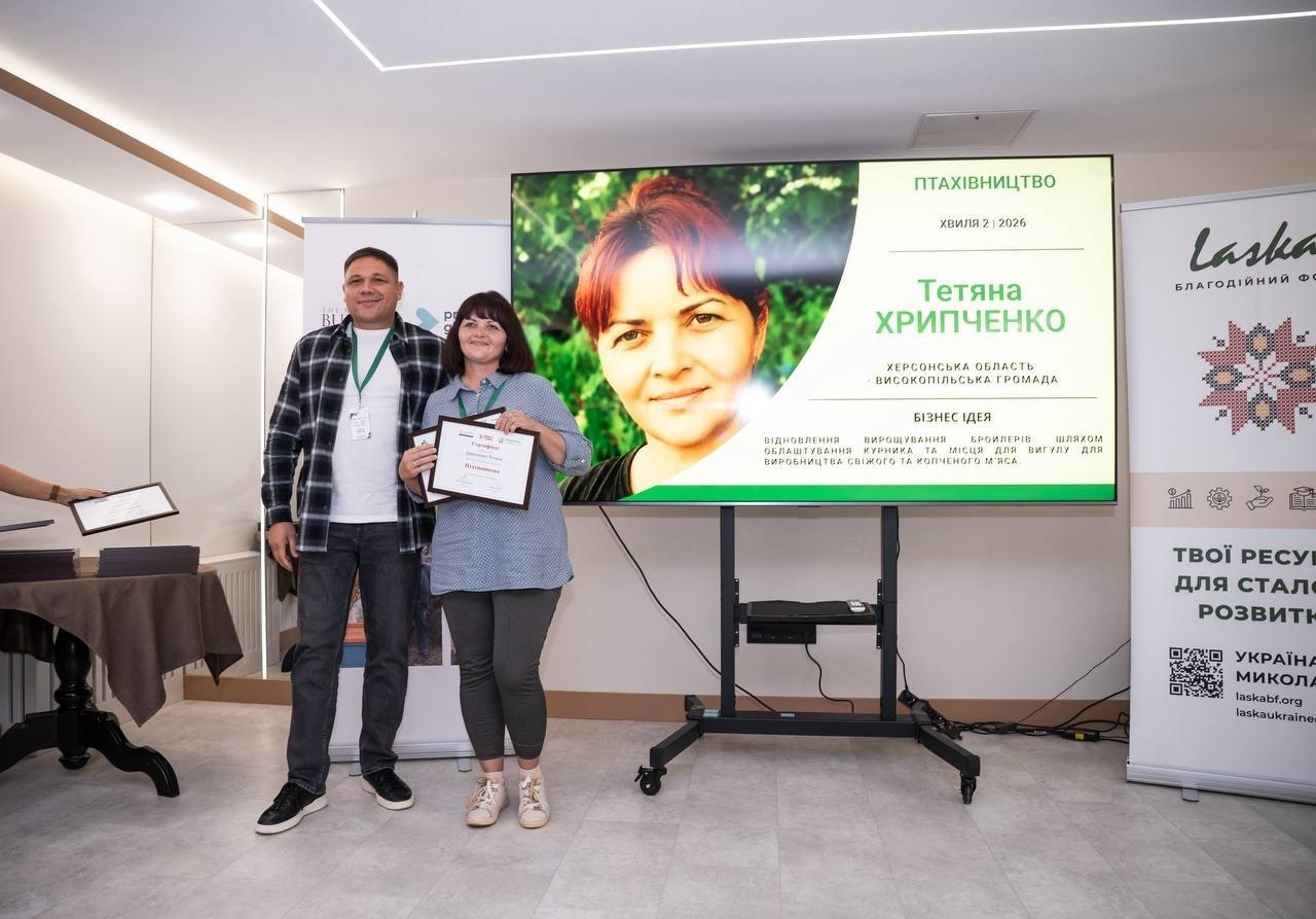 На світлині показано момент вручення грантового сертифіката фермерці Тетяні Хрипченко з Високопільської громади Херсонської області під час форуму в Миколаєві. Ліворуч біля великого екрана стоїть директор Департаменту розвитку сільського господарства та зрошення Херсонської ОДА Дмитро Юнусов у картатій чорно-білій сорочці та білій футболці, а поруч із ним радісно позує переможниця конкурсу Тетяна Хрипченко в блакитній блузці та сірих лосинах, яка тримає в руках свій офіційний диплом у темній рамці. На великому мультимедійному екрані по центру продемонстровано яскравий слайд презентації з фотокарткою переможниці, великим зеленим написом «Тетяна ХРИПЧЕНКО», зазначенням напряму «ПТАХІВНИЦТВО» та описом її бізнес-ідеї: «ВІДНОВЛЕННЯ ВИРОЩУВАННЯ БРОЙЛЕРІВ ШЛЯХОМ ОБЛАШТУВАННЯ КУРНИКА ТА МІСЦЯ ДЛЯ ВИГУЛУ ДЛЯ ВИРОБНИЦТВА СВІЖОГО ТА КОПЧЕНОГО М'ЯСА». Зліва від екрана помітно частину столу з купою дипломів, а праворуч височіє мобільний банер благодійного фонду «ЛАСКА» із гаслом «ТВОЇ РЕСУРСИ ДЛЯ СТАЛОГО РОЗВИТКУ» та QR-кодом.