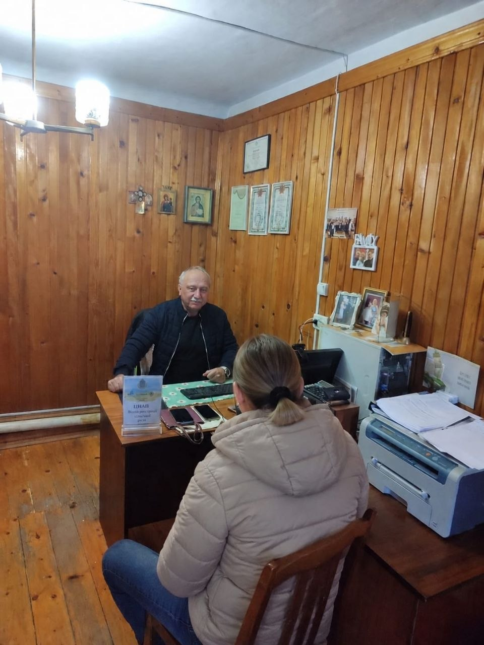 На світлині показано робочий процес надання адміністративних послуг для внутрішньо переміщених осіб із Херсонщини в чернівецькому координаційному центрі «Вільні разом». За робочим дерев'яним столом сидить адміністратор ЦНАП Володимир Чебан — чоловік у чорній куртці на блискавці та чорній кофті, який уважно вислуховує відвідувачку. Перед ним на стільці спиною до об'єктива сидить жінка у світло-бежевій куртці з капюшоном та синіх джинсах, волосся якої зібране у хвіст. На столі перед адміністратором встановлено невелику табличку з офіційним гербом та написом «ЦНАП», а поруч лежать мобільні телефони й комп'ютерна клавіатура на зеленому килимку. Робочий кабінет повністю оздоблений дерев'яною вагонкою на стінах та підлозі, де на задньому плані над комодом із принтером та документами розвішано офіційні грамоти у рамках, подяки та сімейні фотокартки.