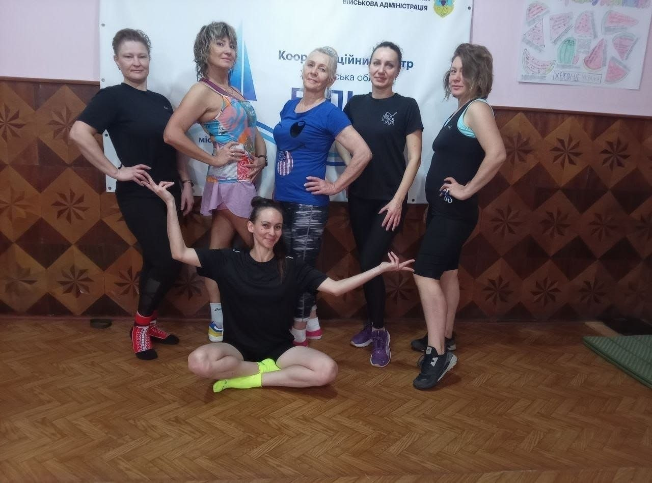 На світлині показано учасниць розважально-оздоровчого фітнес-тренування «Будь сильною» з Іриною Котовою, організованого для переселенок із Херсонщини в чернівецькому хабі «Вільні разом». У залі з дерев'яним паркетом та оригінальними коричневими стінами зі шпалерами у великий ромбовидний орнамент для спільного фото ефектно позують шість усміхнених жінок у спортивній формі: на передньому плані по центру на підлозі сидить у глибокому присяді тренера в чорній футболці, чорних шортах та яскраво-салатових шкарпетках, яка граційно розвела руки в боки; за нею вишикувалися ще п'ять учасниць, які тримають руки на талії або елегантно відвели їх убік. Зліва направо у верхньому ряду стоять: жінка в чорній футболці, лосинах та яскраво-червоних шкарпетках; жінка у різнокольоровій майці та бузкових шортах; по центру — жінка з короткою зачіскою у синій фірмовій футболці з американським патріотичним принтом та темних лосинах; далі — дівчина у чорному спортивному костюмі та фіолетових кросівках, а крайньою праворуч — жінка в чорному топі, чорних шортах та кросівках «Puma». Група позує на тлі великого білого банера координаційного центру, де вгорі помітно герб і напис «ВІЙСЬКОВА АДМІНІСТРАЦІЯ», по центру виведено текст «Координаційний центр Херсонська область Вільні разом», а праворуч на рожевій стіні закріплено малюнок із зображенням часточок кавуна та патріотичним написом «Херсонщина».