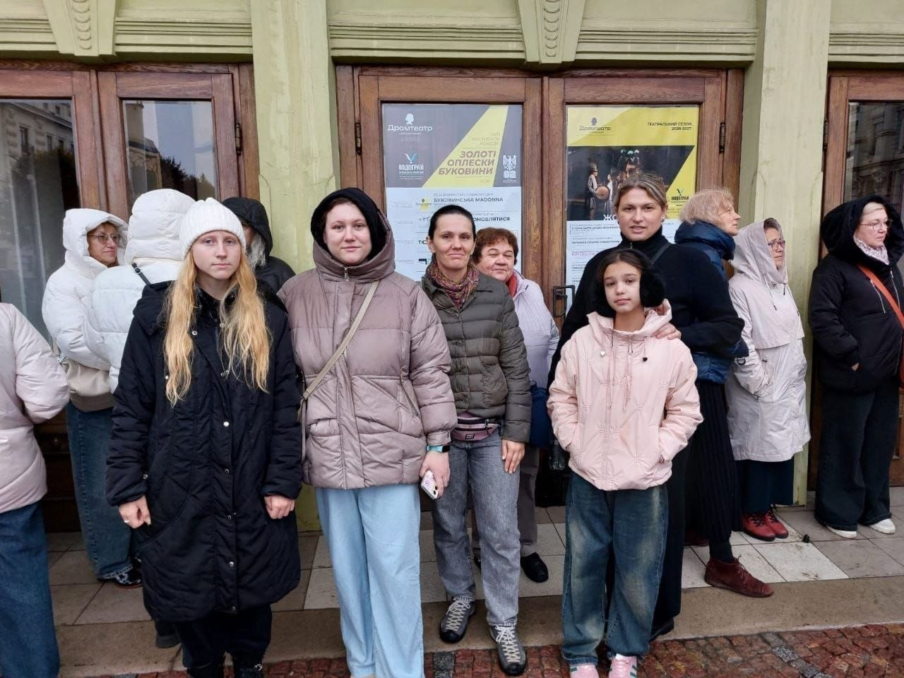 На світлині показано групу херсонських переселенців перед входом до Чернівецького музично-драматичного театру ім. Ольги Кобилянської перед переглядом вистави «Ромео і Джульєтта». На передньому плані на тлі дерев'яних дверей театру вишикувалися глядачі в теплому осінньому одязі, серед яких на першому плані ліворуч стоїть дівчина у довгій чорній куртці та білій в'язаній шапці, поруч із нею — дівчина у коричневій дутій куртці та світло-блакитних широких джинсах. Безпосередньо між нею та дівчинкою в рожевій куртці по центру стоїть жінка в темній куртці та строгатому коричнево-білому шарфі, яка посміхається в об'єктив, а праворуч позує ще одна жінка, яка обіймає за плечі згадану дівчинку в рожевій куртці та широких джинсах. На скляних панелях великих дерев'яних дверей позаду присутніх наклеєно великі офіційні театральні афіші з написами «Драмтеатр», емблемою фестивалю «Золоті оплески Буковини» та розкладом найближчих вистав. Праворуч та ліворуч вздовж світло-зеленого фасаду старовинної будівлі театру стоять інші відвідувачі в куртках, пальтах та хустках, які очікують початку культурного заходу.