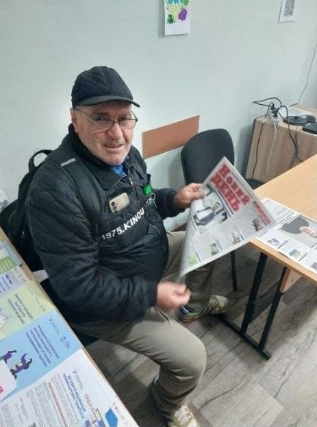 На світлині показано літнього чоловіка-переселенця з Херсонщини, який тримає у руках свіже друковане видання газети «Новий день» у чернівецькому координаційному центрі «Вільні разом». Чоловік в окулярах, одягнений у чорну стьобану куртку з білим написом, бежеві штани, світлі кросівки та чорну кепку, сидить на стільці в офісному приміщенні хабу. Він тримає розгорнуту газету з великим червоним логотипом «НОВИЙ ДЕНЬ», на першій шпальті якої розміщено фотографії та актуальні новини. Поруч із ним розташований довгий дерев'яний стіл світлого відтінку на чорних металевих ніжках, де розкладено інші примірники преси та інформаційні буклети. На передньому плані ліворуч на столі лежать кольорові юридичні та соціальні пам'ятки для внутрішньо переміщених осіб, а на задньому плані помітно світлу стіну з малюнками, комп'ютерні дроти та частину офісних меблів.