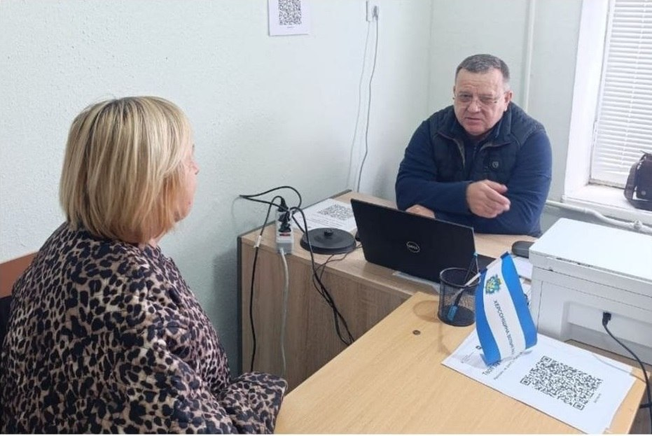 На світлині показано робочий момент особистого прийому громадян, який проводив начальник Виноградівської СВА Валерій Мельниченко для переселенців із Херсонщини в чернівецькому координаційному центрі «Вільні разом». Праворуч за офісним столом сидить керівник військової адміністрації — чоловік в окулярах, одягнений у синю кофту та темний стьобаний жилет, який активно спілкується з відвідувачкою та жестикулює правою рукою. Перед ним на столі встановлено відкритий чорний ноутбук, а поруч лежать документи та стоїть мережевий подовжувач. На передньому плані ліворуч спиною до об'єктива сидить жінка-відвідувачка у кофті з леопардовим принтом, яка прийшла на консультацію. На передньому кутовому столі перед нею розміщено настільний біло-синій прапорець із гербом та написом «ХЕРСОНЩИНА ВІЛЬНІ РАЗОМ», під яким лежать друковані інформаційні листи з великим QR-кодом для переходу на електронні ресурси. Прийом проходить у світлому кабінеті, де на задньому плані праворуч помітно велике вікно із закритими білими горизонтальними жалюзі та великий білий БФП-принтер на тумбі.