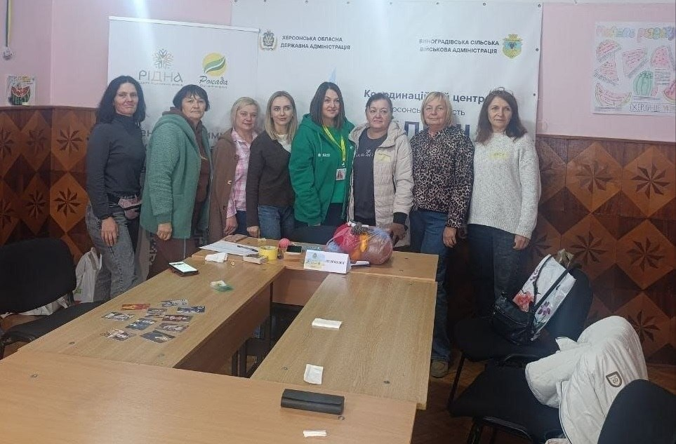 На світлині показано учасниць та організаторів тренінгу із профілактики емоційного вигорання в межах проєкту «Жіноче лідерство» для переселенок із Херсонщини в чернівецькому хабі «Вільні разом». У залі з оригінальним дерев'яним облицюванням стін у ромбовидний візерунок вишикувалися в один ряд вісім жінок, які позують навколо зсунутих літерою «П» світло-дерев'яних столів. У центрі групи стоїть фахівчиня у фірмовому зеленому худі БФ «Рокада» із білим ланьярдом на шиї. Інші учасниці одягнені в теплий повсякденний одяг: кофти, куртки, кардигани та светри. На столах перед ними розкладено метафоричні асоціативні картки, канцелярію, встановлено табличку «ПСИХОЛОГ», а також лежить великий пакунок із нитками та матеріалами для рукоділля. На задньому плані кадру розгорнуто великий білий офіційний банер, на якому чітко видно написи та логотипи: «ХЕРСОНСЬКА ОБЛАСНА ДЕРЖАВНА АДМІНІСТРАЦІЯ», «ВИНОГРАДІВСЬКА СІЛЬСЬКА ВІЙСЬКОВА АДМІНІСТРАЦІЯ» та «Координаційний центр Вільні разом», а ліворуч на стіні помітно банер ініціативи «РІДНА ЦЕНТР ПІДТРИМКИ ЖІНОК» та БФ «Рокада».