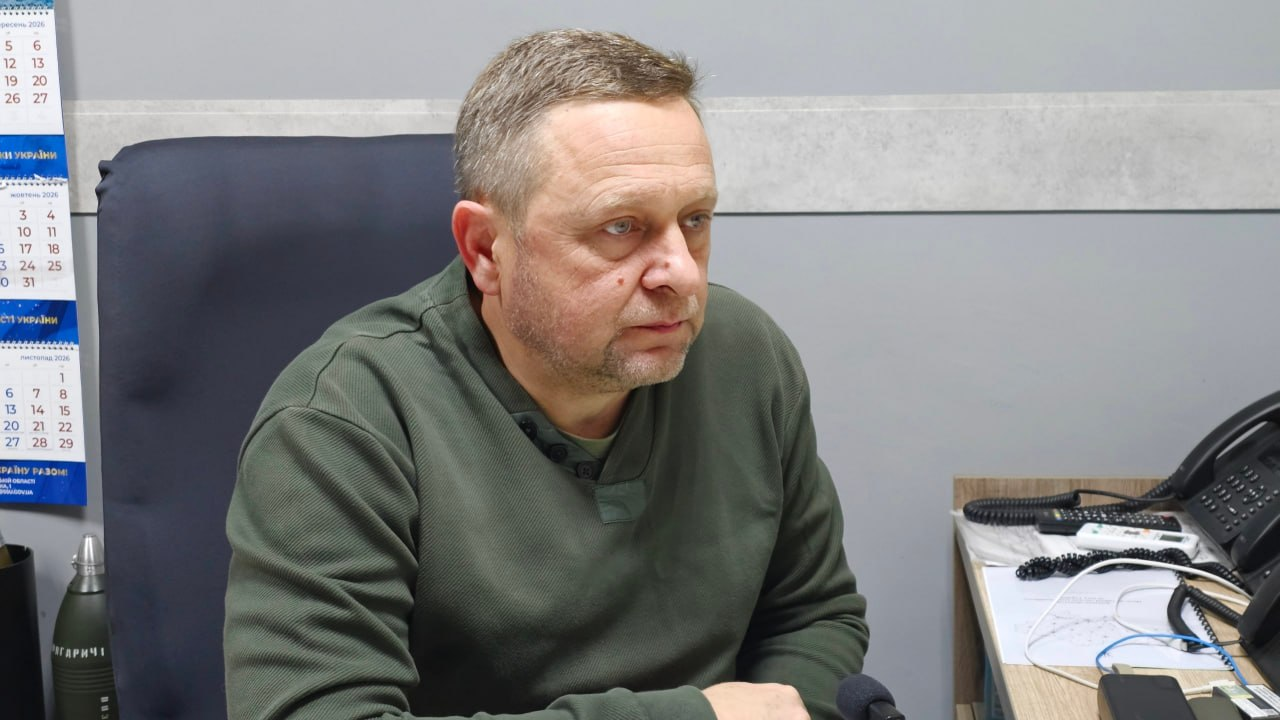 На світлині зображено першого заступника начальника Херсонської ОВА Дмитра Бутрія під час засідання обласної робочої групи «Прозорість і підзвітність». Чоловік середнього віку з короткою сивіючою стрижкою та легкою щетиною, одягнений у мілітарі-пуловер кольору хакі на ґудзиках, сидить у великому чорному офісному кріслі та зосереджено дивиться перед собою. Подія розгортається у кабінеті на тлі сірих стін, де ліворуч на стіні закріплено вертикальний настінний тримісячний календар на жовтень 2026 року. Під календарем на столі помітно сувенірний макет бойового снаряда-гільзи з патріотичним написом, а праворуч на робочому столі розташовані стаціонарний офісний телефон із купою кнопок, слухавкою та проводами, пульт дистанційного керування, біла слухавка радіотелефона та паперові документи з графіками й роздрукованими картами.