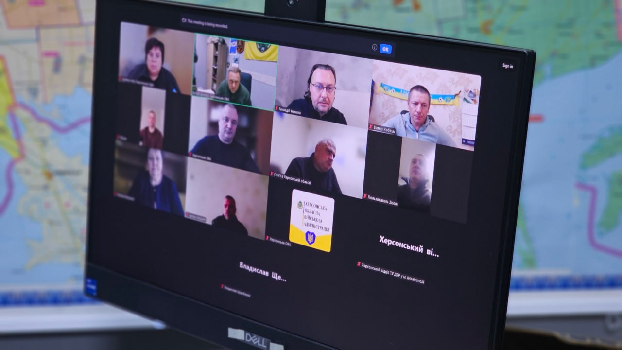 На світлині крупним планом показано екран комп'ютерного монітора під час онлайн-засідання обласної робочої групи «Прозорість і підзвітність». На дисплеї запущено груповий відеодзвінок у застосунку Zoom, де у віконцях відображаються учасники наради, які долучилися до обговорення дистанційно. У другому віконці верхнього ряду ліворуч зафіксовано першого заступника начальника Херсонської ОВА Дмитра Бутрія в мілітарі-пуловері кольору хакі на тлі кабінету з настінним календарем. Серед інших спікерів у віконцях присутні керівники Департаментів та представники правоохоронних органів, один з яких виходить на зв'язок на тлі Державного Прапора України, а в окремому віконці внизу виведено офіційний білий логотип із гербом та написом «ХЕРСОНСЬКА ОБЛАСНА ВІЙСЬКОВА АДМІНІСТРАЦІЯ». На задньому плані кадру позаду монітора видніється велика настінна топографічна карта Херсонської області.