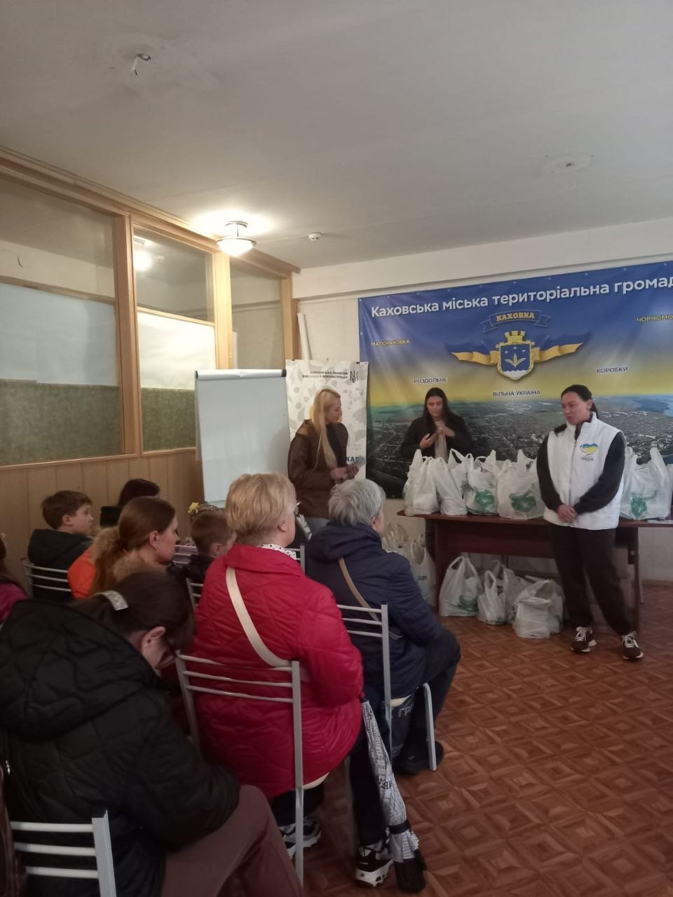 На світлині зафіксовано процес видачі гуманітарної допомоги родинам переселенців із Каховської громади, організований за сприяння БФ «Надія» у київському координаційному центрі «Вільні разом». У залі засідань на стільцях сидить група херсонців у верхньому одязі, які уважно слухають організаторів. Біля довгого столу, повністю заставленого зібраними білими пакетами з продуктовими наборами, стоять три жінки: праворуч перед присутніми виступає волонтерка у білому фірмовому жилеті з логотипом «НАДІЯ БФ», по центру за столом перебуває друга фахівчиня у чорній кофті, а ліворуч біля мобільного фліпчарта — третя представниця у коричневій куртці. Захід проходить на тлі великого синього офіційного банера з картою місцевості, золотим гербом та написом «Каховська міська територіальна громада», де зазначено назви населених пунктів: «КАХОВКА», «МАЛОКАХОВКА», «РОЗДОЛЬНЕ», «ВІЛЬНА УКРАЇНА», «КОРОБКИ» та «ЧОРНОМОРІВКА».