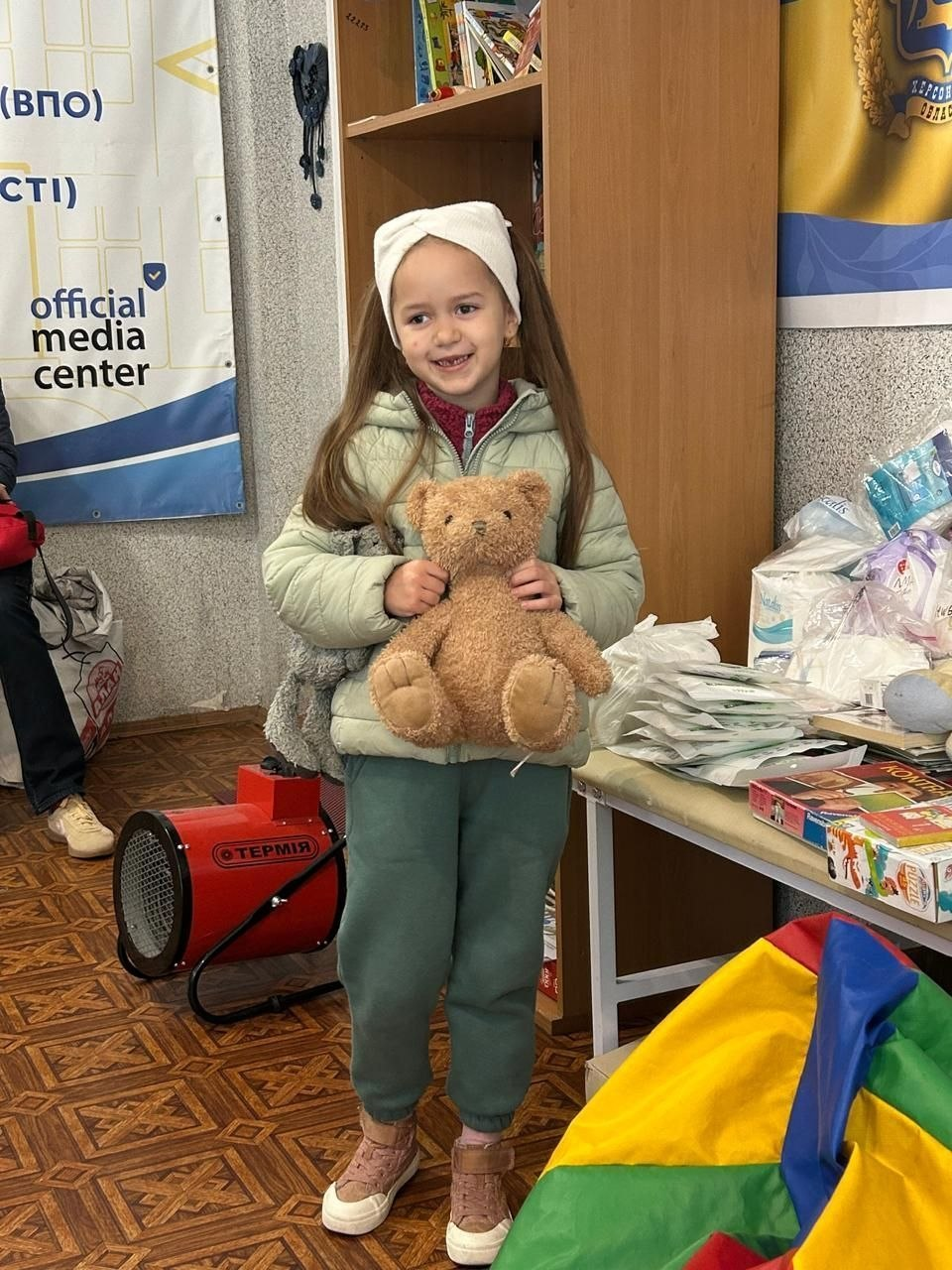 На світлині зображено маленьку херсонську переселенку, яка радісно посміхається в ігровій кімнаті київського координаційного центру «Вільні разом». Дівчинка з довгим русявим волоссям, одягнена у світлу білу пов'язку на голові, світло-зелену дуту куртку, зелені спортивні штани та бежеві кросівки, міцно обіймає обома руками свого нового плюшевого ведмедика коричневого кольору. Вона стоїть посеред дитячого куточка на тлі дерев'яної шафи-стелажа з дитячими книжками. Праворуч розташовано стіл, на якому розкладено набори засобів гігієни, підгузки та настільні ігри з пазлами, а на підлозі перед ним лежить яскравий різнокольоровий пуф-мішок. Ліворуч на підлозі встановлено червону електричну теплову гармату з чорним написом «ТЕРМІЯ», а на задньому плані видно частину інформаційного банера з англійським написом «official media center» та герб Херсонської області на правому банері.
