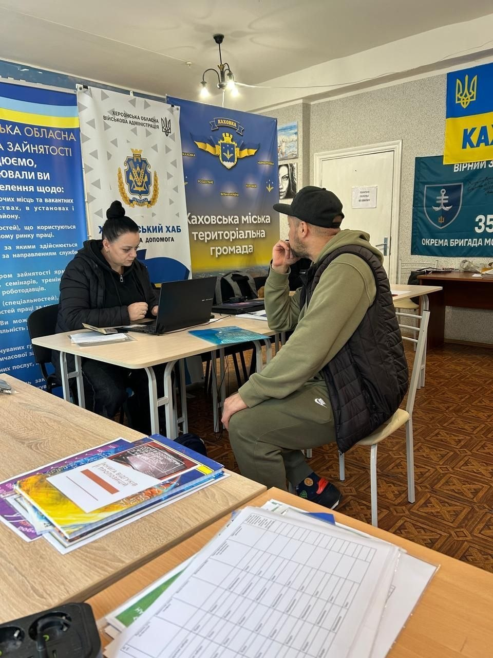 На світлині показано робочий момент надання фахових юридичних консультацій для внутрішньо переміщених осіб із Херсонщини в київському координаційному центрі «Вільні разом». Ліворуч за світлим робочим столом сидить жінка-юристка з темним волоссям, зібраним у пучок, яка одягнена в чорну куртку та зосереджено вносить дані у відкритий чорний ноутбук. Навпроти неї на стільці в профіль сидить чоловік-відвідувач у зеленому спортивному костюмі, чорному стьобаному жилеті та чорній кепці, який уважно слухає роз'яснення фахівця. На передньому плані на столах розкладено папки з друкованими інформаційними матеріалами, буклети та чисті бланки заяв для заповнення. Прийом громадян проходить на тлі мобільних банерів Херсонської ОВА «Херсонський хаб. Інформація та допомога», Каховської міської територіальної громади та синього банера Державної служби зайнятості, а праворуч на стіні кабінету майорить темно-синій прапор 35-ї окремої бригади морської піхоти поруч із Державним прапором України.