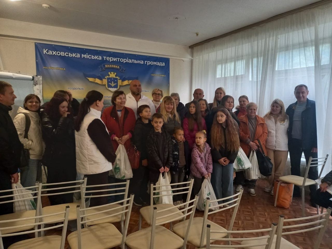 На світлині показано велику групу переселенців із Каховської громади разом із волонтерами під час видачі продуктових наборів у київському координаційному центрі «Вільні разом». У залі засідань для спільного фото зібралися дорослі, літні люди та діти, які тримають у руках отримані великі білі пакети з гуманітарною допомогою. У передньому ряду ліворуч стоїть волонтерка у білому фірмовому жилеті з логотипом «НАДІЯ БФ», обернена боком до об'єктива, а перед нею та праворуч вишикувалися дітлахи різного віку та матусі в осінніх куртках і пальтах. Серед присутніх у центрі заднього ряду виділяється кремезний чоловік у білій спортивній кофті, а крайнім праворуч стоїть чоловік у синій куртці та блакитному светрі. На передньому плані видно порожні світлі стільці, розставлені рядами, а вся група позує на тлі великого синього офіційного банера з картою місцевості, золотим гербом та написом «Каховська міська територіальна громада».