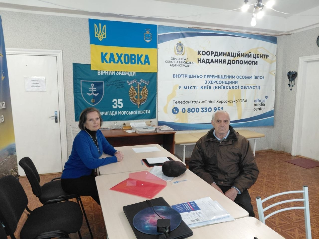На світлині показано робочий момент надання фахових юридичних консультацій для внутрішньо переміщених осіб із Херсонщини в київському координаційному центрі надання допомоги «Вільні разом». У кабінеті з лінолеумом із геометричним візерунком за великим світлим кутовим столом сидять двоє людей: ліворуч — жінка-юристка в яскраво-синій кофті та чорних штанях, яка консультує відвідувача, а праворуч навпроти неї — літній чоловік у коричневій зимовій куртці з хутряним коміром, який уважно слухає роз'яснення. На столі перед ними розкладено прозору папку з червоним клапаном, чисті аркуші паперу для нотаток та великий синьо-білий друкований інформаційний буклет із правовими роз'ясненнями, а поруч лежить круглий килимок із космічним принтом. На задньому плані кадру на стіні розвішано офіційну символіку: великий білий банер із написом «КООРДИНАЦІЙНИЙ ЦЕНТР НАДАННЯ ДОПОМОГИ ВНУТРІШНЬО ПЕРЕМІЩЕНИМ ОСОБАМ (ВПО) З ХЕРСОНЩИНИ У МІСТІ КИЇВ» та телефоном гарячої лінії, жовто-блакитний прапор із написом «КАХОВКА» та темно-синій прапор 35-ї окремої бригади морської піхоти.