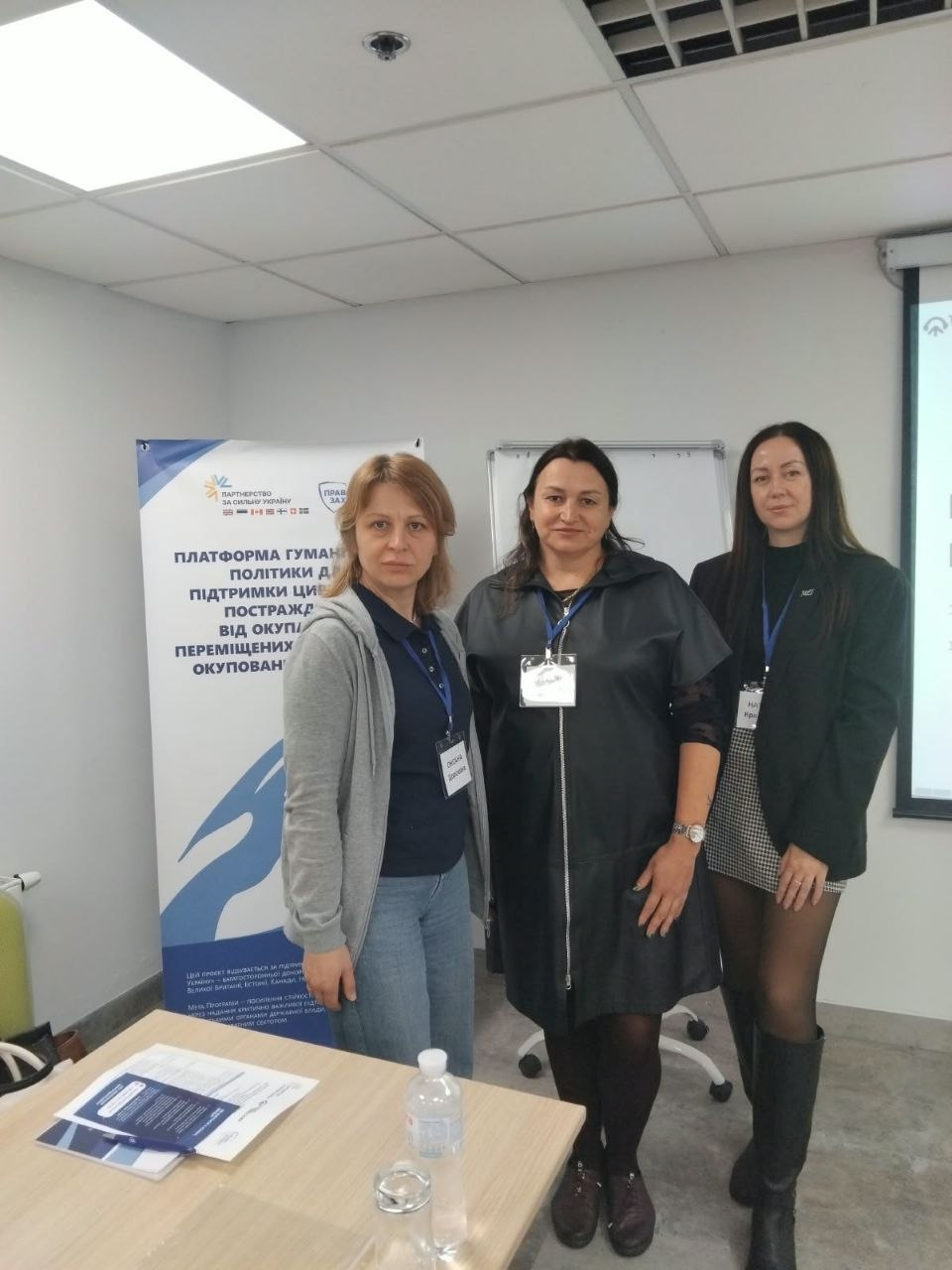 На світлині показано представниць та юристок під час надання правової допомоги для внутрішньо переміщених осіб із Херсонщини в межах діяльності київського координаційного центру «Вільні разом». У світлому сучасному кабінеті з підвісною стелею стоять три жінки-фахівчині, на шиях яких закріплено сині стрічки-ланьярд з акредитаційними бейджами учасників заходу. Ліворуч стоїть жінка зі світло-русявим волоссям у сірій кофті на блискавці, темно-синій футболці-поло та блакитних джинсах. По центру позує жінка з темним волоссям у стильному довгому чорному шкіряному кардигані-плащі на блискавці та темних колготках, а праворуч — довгокоса представниця у чорному класичному піджаку, короткій картатій спідниці та високих чорних шкіряних чоботах. На задньому плані ліворуч височіє мобільний ролап-банер із великим написом «ПЛАТФОРМА ГУМАНІТАРНОЇ ПОЛІТИКИ ДЛЯ ПІДТРИМКИ ЦИВІЛЬНОГО НАСЕЛЕННЯ, ПОСТРАЖДАЛОГО ВІД ОКУПАЦІЇ ТА ПЕРЕМІЩЕНИХ З ТИМЧАСОВО ОКУПОВАНИХ ТЕРИТОРІЙ», поруч розташована біла маркерна дошка-фліпчарт, а на столі на передньому плані розкладено друковані інформаційні брошури про права ВПО, стоїть пляшка води та склянка.