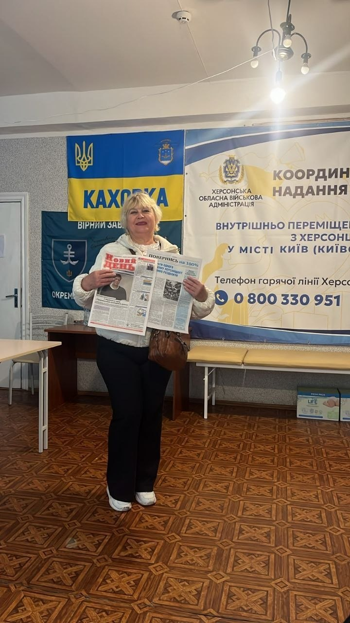 На світлині показано жінку-переселенку з Херсонщини у київському координаційному центрі надання допомоги «Вільні разом», яка тримає в руках друковані видання. У центрі кадру в повний зріст стоїть усміхнена жінка з короткою світлою зачіскою, одягнена у світлу теплу куртку, чорні штани та білі кросівки, з коричневою сумочкою через плече. Вона гордо демонструє розгорнуту свіжу газету, де зліва чітко видно великий червоний логотип видання «НОВИЙ ДЕНЬ», а на правій сторінці надруковано назву спецвипуску «ПОВЕРНИСЬ НА 100%» з фотоматеріалами та статтями. Подія розгортається у кабінеті з лінолеумом із коричневим геометричним візерунком, де на задньому плані розвішано офіційну символіку: великий білий банер із написом «ХЕРСОНСЬКА ОБЛАСНА ВІЙСЬКОВА АДМІНІСТРАЦІЯ. КООРДИНАЦІЙНИЙ ЦЕНТР НАДАННЯ ДОПОМОГИ ВНУТРІШНЬО ПЕРЕМІЩЕНИМ ОСОБАМ З ХЕРСОНЩИНИ У МІСТІ КИЇВ» та телефоном гарячої лінії «0 800 330 951», жовто-блакитний прапор із написом «КАХОВКА» та темно-синій прапор 35-ї окремої бригади морської піхоти.