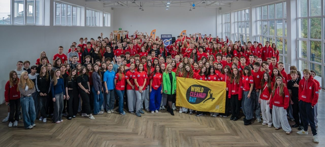 На світлині показано масштабне спільне фото чисельних учасників проєкту розвитку талановитої молоді «Залізна Зміна», серед яких дванадцять дітей із Херсонщини. У центрі великої та світлої спортивної зали з високими вікнами та дерев'яним паркетом зібралася величезна група підлітків, більшість із яких одягнені в однакові фірмові червоні худі чи футболки з білими написами. У самому центрі першого ряду стоїть координаторка або почесна гостя у яскравому салатовому піджаку та чорних штанях, а поруч із нею діти тримають великий жовтий прапор із символікою всесвітнього дня прибирання «WORLD CLEANUP DAY». На передньому плані ліворуч виділяються дівчата у повсякденному одязі (широких синіх джинсах, чорній кофті, синій та блакитній футболках), а праворуч позують підлітки у спортивних костюмах та худі. На задньому плані в глибині залу помітно фірмові мобільні банери та синій прапор із круглим логотипом проєкту «ЗАЛІЗНА ЗМІНА».