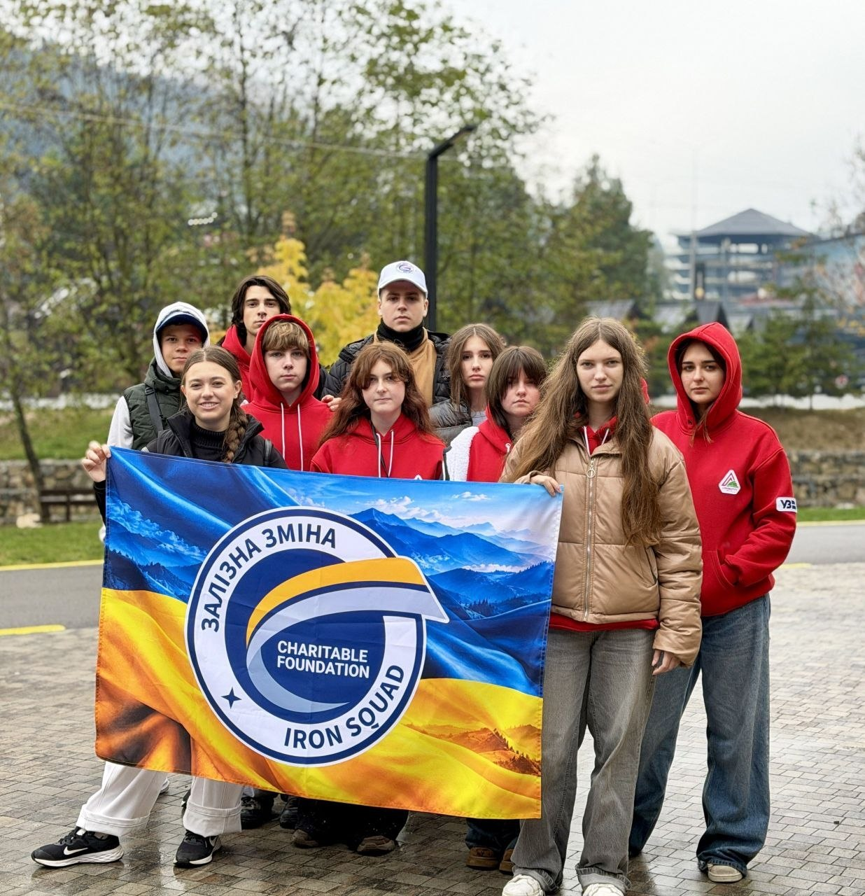 На світлині показано групу херсонських учасників проєкту розвитку талановитої молоді «Залізна Зміна» під час поїздки на Прикарпаття. Одинадцять підлітків вишикувалися в кілька рядів на брукованому майданчику просто неба і разом тримають великий синьо-жовтий прапор із гірським пейзажем, фірмовим круглим логотипом та написами: «ЗАЛІЗНА ЗМІНА. CHARITABLE FOUNDATION IRON SQUAD». Більшість юнаків та дівчат одягнені в однакові фірмові червоні худі з білим трикутним логотипом на грудях. На передньому плані ліворуч дівчина з косою у чорній безрукавці посміхається в об'єктив, по центру стоїть дівчина у червоному худі, а праворуч позують довгокоса дівчина в бежевій дутій куртці, сірих джинсах та білих кросівках і дівчина в червоному худі та широких синіх джинсах. У задніх рядах видно хлопців та дівчат у куртках, жилетках, худі та кепках, а на задньому плані кадру на тлі хмарного піднебіння височіють зелені ялини, гори, ліхтарні стовпи та заміські дерев'яні будівлі.