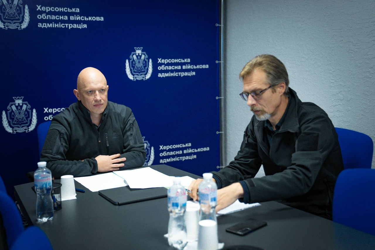 На світлині показано робочий момент засідання Координаційного штабу з питань підтримки евакуйованих жителів за участю представників Херсонщини. За великим чорним офісним столом сидять двоє чоловіків: ліворуч — заступник начальника Херсонської ОВА Євгеній Ігнатенко у темно-сірій тактичній флісовій кофті, який склав руки перед собою, а праворуч навпроти нього — заступник директора Департаменту з питань цивільного захисту та оборонної роботи Херсонської ОДА Володимир Бєляєв в окулярах та чорній куртці, який зосереджено робить нотатки кульковою ручкою на білих аркушах паперу. На столі перед ними розкладено робочі документи, планшети, смартфони, стоять пластикові пляшки з водою та паперові стаканчики. Зустріч проходить у залі засідань на тлі великого темно-синього офіційного банера, на якому нанесено герби та численні білі написи «Херсонська обласна військова адміністрація».