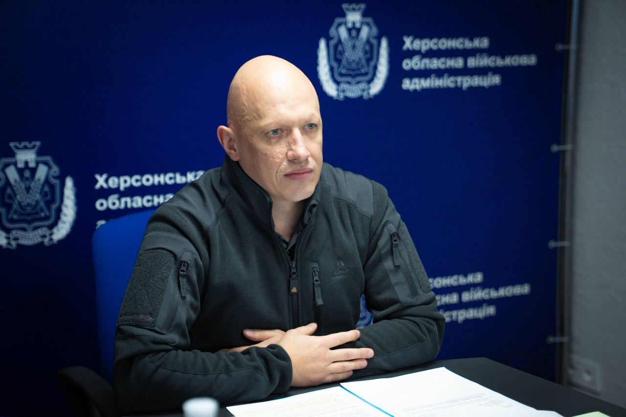 На світлині зображено заступника начальника Херсонської ОВА Євгенія Ігнатенка під час онлайн-засідання Координаційного штабу з питань підтримки евакуйованих жителів. Він сидить у блакитному офісному кріслі за великим темним столом, одягнений у темно-сіру тактичну флісову кофту на блискавці із велкро-панелями (липучками) та кишенями на рукавах. Чоловік зосереджено слухає інших учасників наради та дивиться вперед, зчепивши пальці рук перед собою на столі поверх розгорнутої білої папки з робочими документами та роздрукованими листами. Зустріч проходить у залі засідань адміністрації на тлі великого офіційного темно-синього банера, на якому нанесено білі круглі герби та численні написи «Херсонська обласна військова адміністрація».