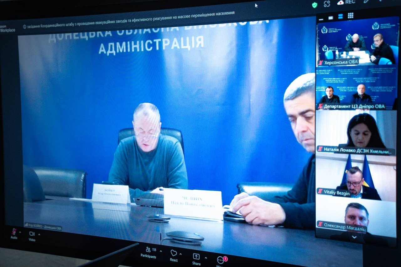 На світлині крупним планом показано великий екран монітора під час онлайн-засідання Координаційного штабу з проведення евакуаційних заходів. У лівій частині екрана на великому головному вікні транслюється включення колег із Донеччини, де на тлі синього банера з написом «ДОНЕЦЬКА ОБЛАСНА ДЕРЖАВНА АДМІНІСТРАЦІЯ» за столом із табличками сидять двоє представників. У правій частині монітора серед віконець інших учасників цифрової наради у першому віконці зверху з підписом «Херсонська ОВА» за робочим столом сидить керівництво Херсонщини — заступник начальня ОВА Євгеній Ігнатенко та заступник директора Департаменту з питань цивільного захисту та оборонної роботи Володимир Бєляєв. Також на екрані відображається віконце з підписом «Vitaliy Bezgin», де на тлі Державного Прапора України виступає Міністр у справах громад, територій та внутрішньо переміщених осіб України Віталій Безгін. У верхній лівій частині інтерфейсу програми Zoom чітко видно тему наради: «засідання Координаційного штабу з проведення евакуаційних заходів та ефективного реагування на масове переміщення населення».