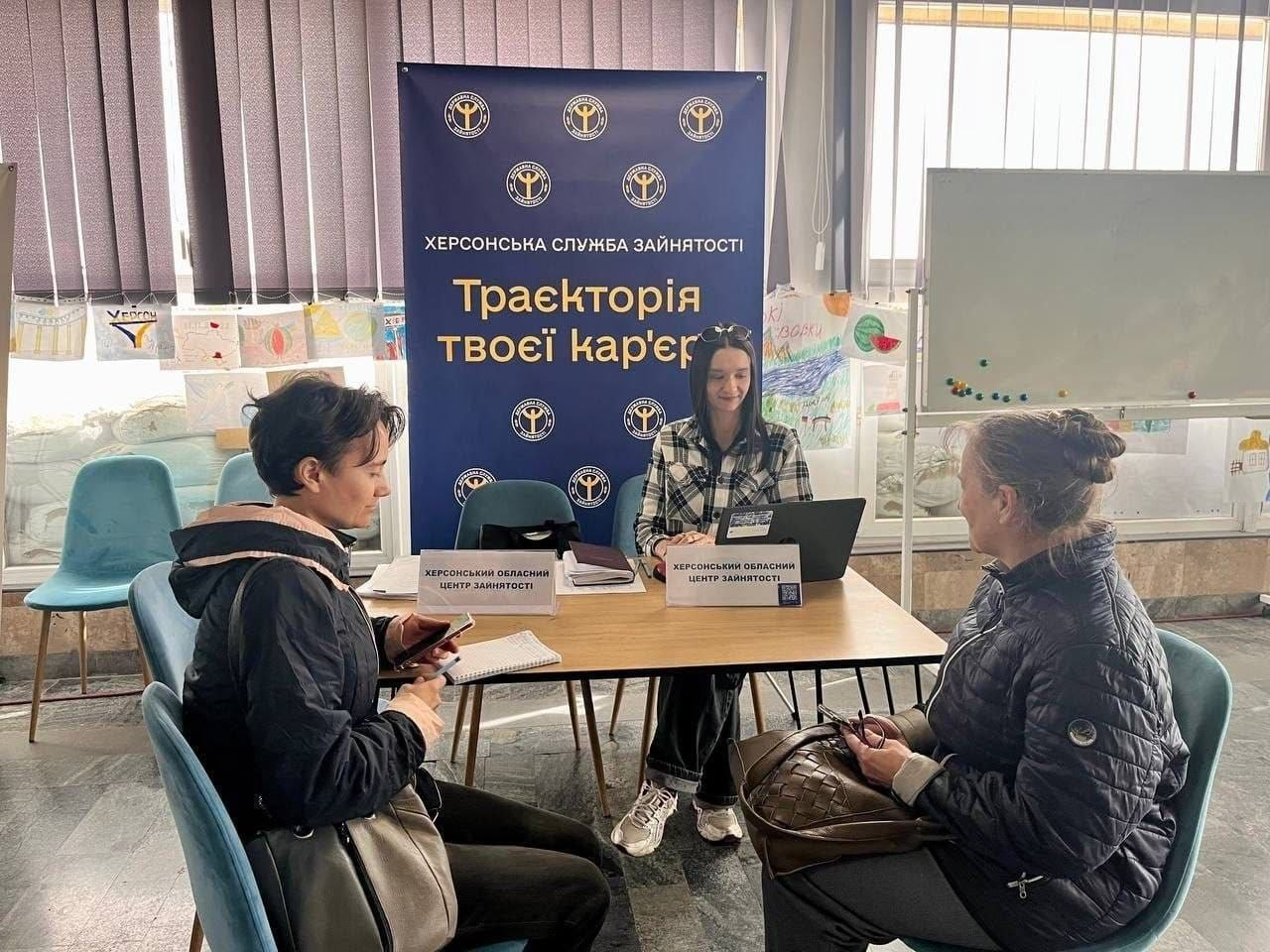 На світлині показано процес надання кар'єрних консультацій для переселенців із Херсонщини в миколаївському координаційному центрі «Вільні разом» фахівцями Херсонської служби зайнятості. За великим дерев'яним столом по центру сидить молода дівчина-консультант у картатій сорочці, яка працює за відкритим чорним ноутбуком, а перед нею на столі встановлено дві білі таблички з написом «ХЕРСОНСЬКИЙ ОБЛАСНИЙ ЦЕНТР ЗАЙНЯТОСТІ». Перед столом на синіх м'яких стільцях сидять дві відвідувачки в темних осінніх куртках, які звернулися по допомогу у працевлаштуванні та тримають у руках свої смартфони й документи. Консультування проходить на тлі великого темно-синього офіційного банера з фірмовими логотипами, написом «ХЕРСОНСЬКА СЛУЖБА ЗАЙНЯТОСТІ» та гаслом «Траєкторія твоєї кар'єри». На задньому плані кадру вздовж великих вікон із вертикальними жалюзі розміщено виставку дитячих малюнків, де помітно зображення кавуна та патріотичні мотиви, а праворуч у кабінеті встановлено велику білу маркерну дошку-фліпчарт.