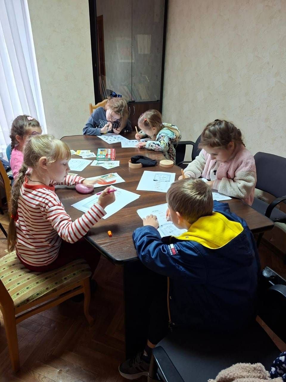 На світлині показано творче та розвивальне заняття для дітей-переселенців із Херсонщини в дитячому просторі миколаївського координаційного центру «Вільні разом». Навколо великого овального дерев'яного столу по центру кімнати сидять семеро малюків, які зосереджено виконують завдання на паперових аркушах, малюють і розфарбовують олівцями. На передньому плані ліворуч сидить світловолоса дівчинка у смугастій червоно-білій кофті з довгим рукавом, тримаючи в руці фломастер, а праворуч навпроти неї зафіксовано хлопчика у темно-синій куртці з яскравим жовтим капюшоном «Tommy Hilfiger». Далі вздовж столу працюють інші дітлахи, серед яких дівчинка у світло-рожевій пухнастій кофті, маленька дитина у світлій куртці з малюнками та підліток у сірому худі з написом «PROUD». На столі перед ними розкладено навчальні роздруківки, коробки з олівцями, кольорові картки та круглі дерев'яні підставки. Заняття проходить у кабінеті з паркетною підлогою та світло-бежевими стінами, де ліворуч велике вікно завішене довгою білою тюлю, а на задньому плані видніється висока темна шафа для документів і книг.