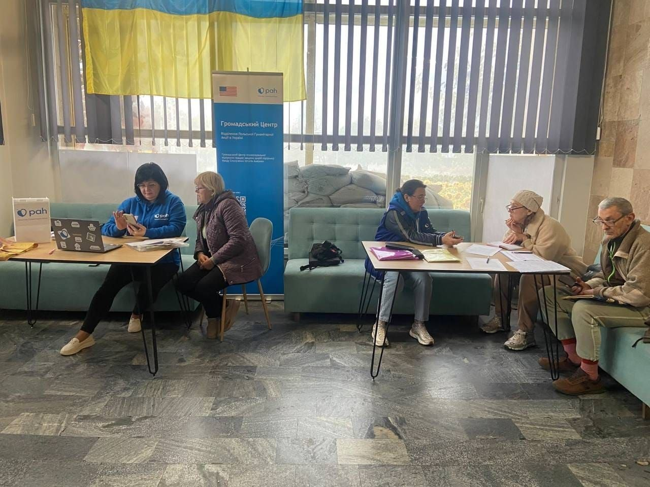 На світлині показано процес реєстрації та надання допомоги для переселенців із Херсонщини в миколаївському координаційному центрі «Вільні разом» від Відділення Польської Гуманітарної Акції в Україні (PAH). У просторій залі за двома окремими столами проходять консультації: ліворуч за першим столом працює фахівчиня у фірмовому синьому худі з логотипом «pah», яка вносить дані у ноутбук, а поруч сидить відвідувачка у бузковій куртці. За другим столом праворуч сидить інша представниця організації у синій жилетці, яка спілкується з літньою жінкою в бежевому пальті та в'язаній світлій шапці й чоловіком у бежевій куртці, окулярах та світло-зелених штанях, що чекають на оформлення гігієнічних наборів. На лівому столі встановлено паперовий фірмовий пакет із логотипом «pah», а по центру кадру височіє синьо-білий мобільний ролап-банер із написом «Громадський Центр. Відділення Польської Гуманітарної Акції в Україні», прапорами США та організації. Захід проходить у приміщенні з мармуровою підлогою та великими вікнами з вертикальними жалюзі, де вгорі закріплено Державний прапор України, а за склом для захисту викладено захисну барикаду з мішків із піском.