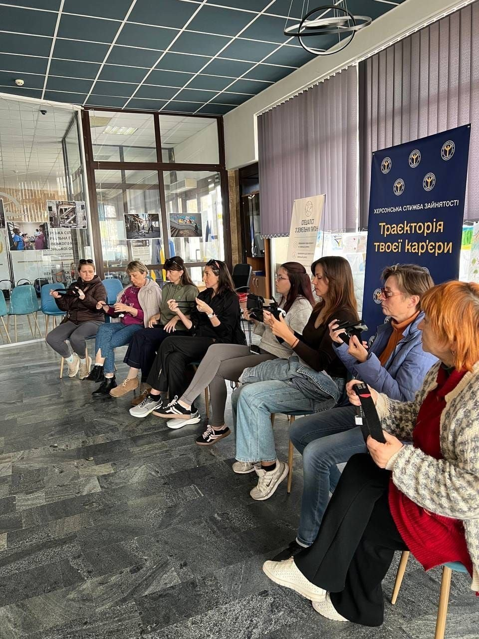 На світлині показано учасниць практичного заняття з домедичної допомоги під час відпрацювання навичок накладання турнікетів у миколаївському координаційному центрі «Вільні разом». У просторій залі з мармуровою підлогою на синіх та дерев'яних стільцях вишикувалися в один напівкруглий ряд група жінок-переселенок із Херсонщини, які зосереджено тримають у руках чорні навчальні медичні джгути-турнікети. Зліва направо в ряду сидять: жінка в чорній куртці та сірих штанях; жінка у фіолетовій кофті та синіх джинсах; жінка у сірій кофті та темних штанях; дівчина в чорній сорочці та сірих лосинах; дівчина у білому світшоті та широких блакитних джинсах; довгокоса дівчина в чорному лонгсліві та синіх джинсах; жінка в окулярах та блакитній куртці, а крайньою праворуч — жінка з яскравим рудим волоссям у світлій в'язаній кофті та червоному кардигані. Праворуч біля них височіє великий темно-синій банер із написом «ХЕРСОНСЬКА СЛУЖБА ЗАЙНЯТОСТІ. Траєкторія твоєї кар'єри», а на задньому плані кадру на тлі великих вікон із вертикальними жалюзі помітно світлий плакат ССО, скляні офісні перегородки з розвішаними фотоматеріалами та сині стільці в іншій частині залу.