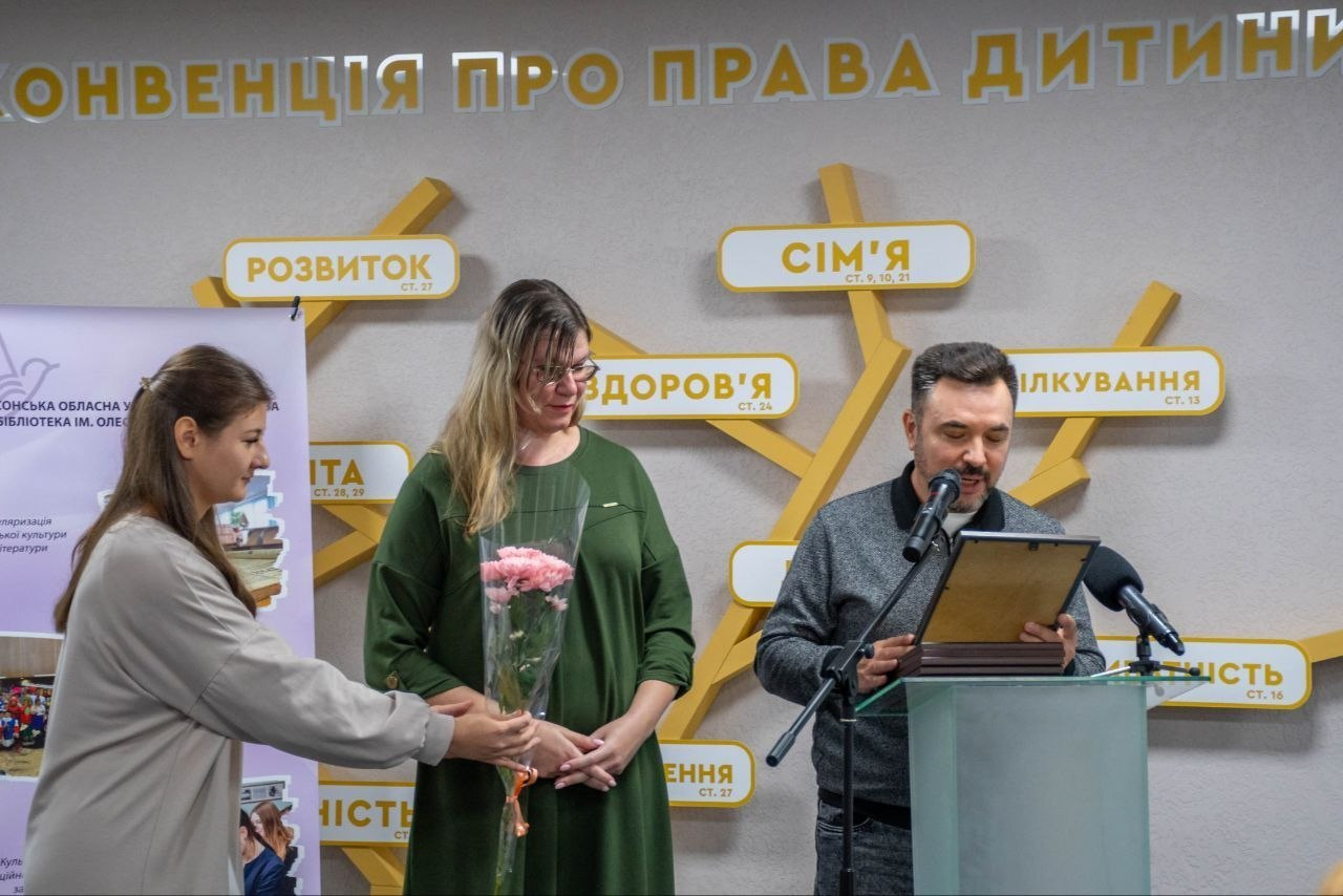 На світлині зафіксовано момент нагородження під час конференції «Бібліотека 4.5.0: адвокація змін» у Кропивницькому, де праворуч за скляною трибуною з мікрофонами стоїть заступник директора Департаменту реалізації гуманітарної політики Херсонської ОДА Андрій Ярьоменко в сірому светрі та зачитує текст почесної грамоти в рамці, яку він тримає в руках. По центру стоїть представниця бібліотечної галузі в окулярах та зеленій сукні, якій ліворуч дівчина у сірій кофті вручає букет рожевих гвоздик у прозорій обгортці. Урочиста подія відбувається на тлі сірої стіни з великим написом угорі «КОНВЕНЦІЯ ПРО ПРАВА ДИТИНИ» та декоративним дерев'яним інсталяційним деревом, на гілках якого розміщено таблички з поняттями «РОЗВИТОК», «СІМ'Я», «ЗДОРОВ'Я», «СПІЛКУВАННЯ» та відповідними номерами статей конвенції, а з лівого краю кадру частково видно інформаційний банер Херсонської обласної універсальної наукової бібліотеки імені Олеся Гончара.