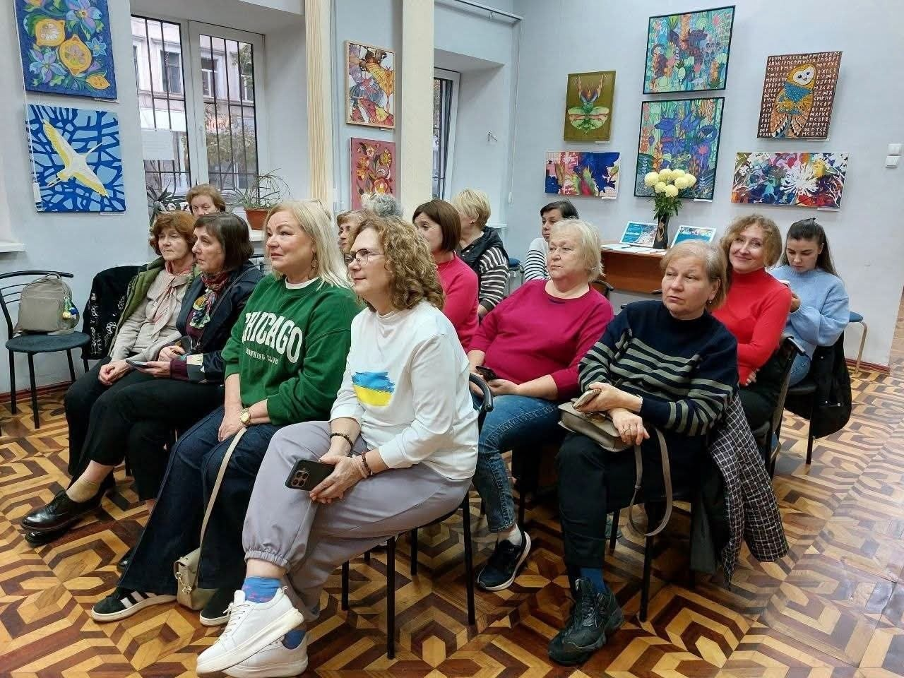 На світлині зображено глядачок творчого заходу для переселенців з Херсонщини в Одесі, які сидять рядами на стільцях у бібліотечній залі та уважно слухають виступ письменника Кирила Булаховського. На передньому плані видно жінок, які були присутні й на загальному фото: одна з них сидить у білому світшоті з патріотичним синьо-жовтим принтом і сірих штанях, тримаючи в руках телефон, а поруч із нею сидить жінка в зеленому світшоті з написом Chicago та темних штанях. За ними та поруч розташовані інші учасниці різного віку в різноколірному повсякденному одязі, зокрема в смугастих кофтах, червоному та малиновому светрах, які зацікавлено дивляться в бік спікера поза кадром. Подія проходить у світлому приміщенні бібліотеки з характерною фігурною дерев'яною паркетною підлогою, а сірі стіни зали прикрашені численними яскравими художніми картинами та дитячими малюнками.