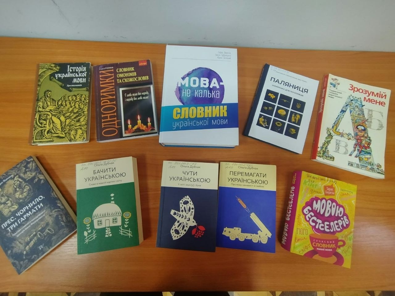 На світлині зображено виставку книг про українську мову на дерев'яному столі під час заходу для переселенців з Херсонщини в Одесі. Книги розкладені у два ряди: у верхньому ряду зліва направо лежать «Історія української мови. Хрестоматія» із зеленою обкладинкою, «Одноримки. Словник омонімів та схожослів», великий біло-синій словник «Мова — не калька», книга «Паляниця. Написано для іноземців» із темно-синіми ілюстраціями та дитяче видання «Зрозумій мене» з літерою «А» на обкладинці. У нижньому ряду зліва направо розміщені історичний роман «Прес, чорнило, три гармати», три патріотичні книги Ольги Дубчак — зелена «Бачити українською», темно-синя «Чути українською» та яскраво-синя «Перемагати українською» із зображенням системи ППО, а замикає ряд яскраво-жовта книга «Мовою бестселерів. Сучасний словник живої мови».