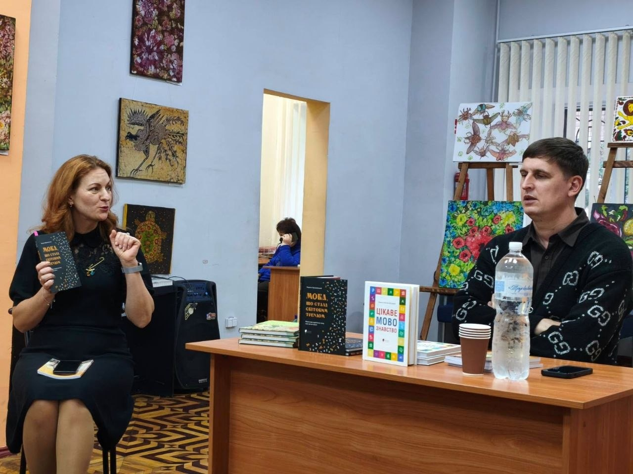 На світлині зображено письменника Кирила Булаховського та модераторку зустрічі під час презентації книги «Мова, що стала світовим трендом» для переселенців з Херсонщини в Одесі. Ліворуч на стільці сиділа жінка-модераторка з довгим рудим волоссям у чорній сукні, яка тримала в руці примірник презентованої книги з темною обкладинкою та емоційно розмовляла, жестикулюючи правою рукою. Праворуч за дерев'яним столом сидів письменник Кирило Булаховський у чорному кардигані з білими фірмовими монограмами Gucci поверх темної сорочки, який уважно слухав або відповідав на запитання. На столі перед ним лежали стоси книг, виставлені примірники видань «Мова, що стала світовим трендом» та «Цікаве мовознавство». Зустріч проходила в бібліотечній залі на тлі світло-бузкових стін із художніми картинами на мольбертах, фігурної паркетної підлоги, а на задньому плані через дверний проріз було видно іншу кімнату, де за столом сиділа працівниця бібліотеки та розмовляла по телефону.
