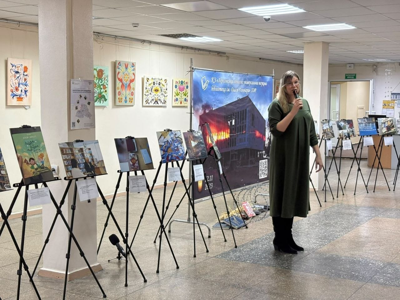 На світлині зображено виступ представниці бібліотечної галузі під час презентації виставкових проєктів на конференції «Бібліотека 4.5.0: адвокація змін» у Кропивницькому. Праворуч на передньому плані стоїть жінка в окулярах, довгій зеленій сукні вільного крою та чорних чоботях, яка тримає в руці мікрофон і виступає перед присутніми. Вона стоїть у просторій залі на тлі великого синього банера КЗ «Херсонська обласна універсальна наукова бібліотека ім. Олеся Гончара» ХОР із фотографією палаючої будівлі, під яким на підлозі розміщено інсталяцію «Коли книга стає мішенню…» з колючим дротом та розсипаними книгами. Ліворуч і праворуч уздовж світлих стін зали розставлено довгі ряди штативів-мольбертів із фотографіями зруйнованих культурних об'єктів у межах виставки «Бібліотеки Херсонщини: злочини росії проти культури України», а на самих стінах вище розвішано яскраві картини та декоративні панно з традиційними українськими народними орнаментами.