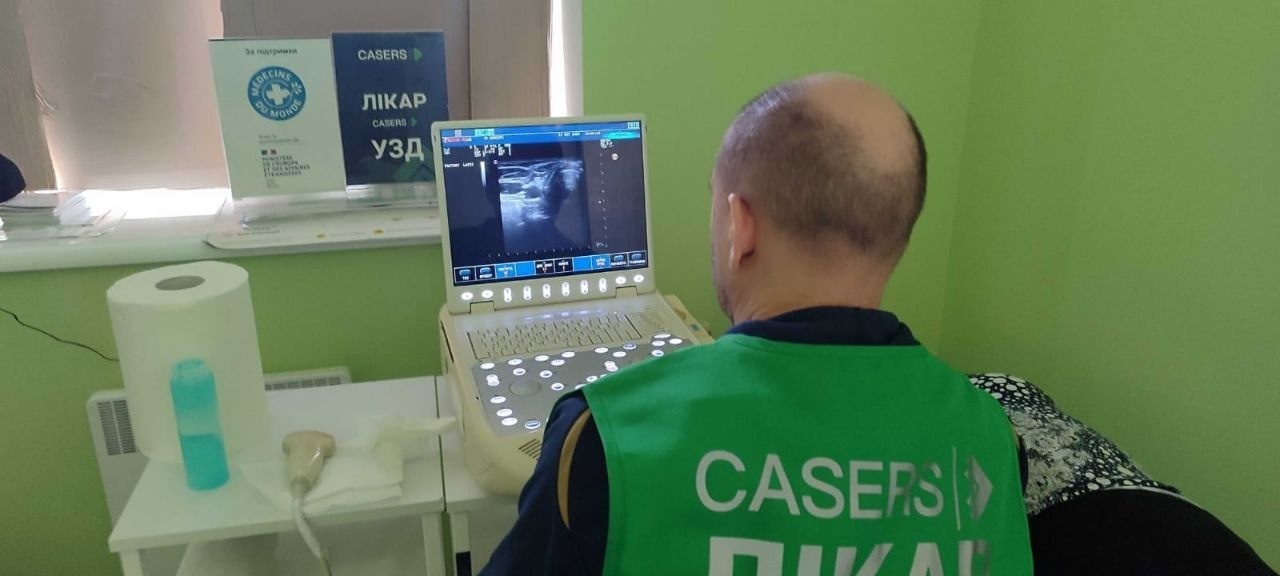 На світлині зафіксовано процес ультразвукової діагностики під час виїзного прийому мобільної медичної бригади для жителів Херсонщини. Спіною до камери сидить лікар у зеленому жилеті з білим написом «CASERS ЛІКАР», який проводить обстеження пацієнта, що лежить на кушетці, та уважно дивиться на екран портативного УЗД-апарата, де відображається чорно-біле діагностичне зображення внутрішніх органів. Обстеження проходить у кабінеті зі світло-зеленими стінами, де ліворуч на маленькому столику розкладено медичні матеріали, зокрема рулон паперових рушників, гель для УЗД у синій пляшечці та датчик апарата, а на підвіконні на задньому плані встановлено прозору інформаційну табличку з синьою наліпкою «CASERS ЛІКАР УЗД» та логотипом міжнародної організації «MÉDECINS DU MONDE» (Лікарі світу).