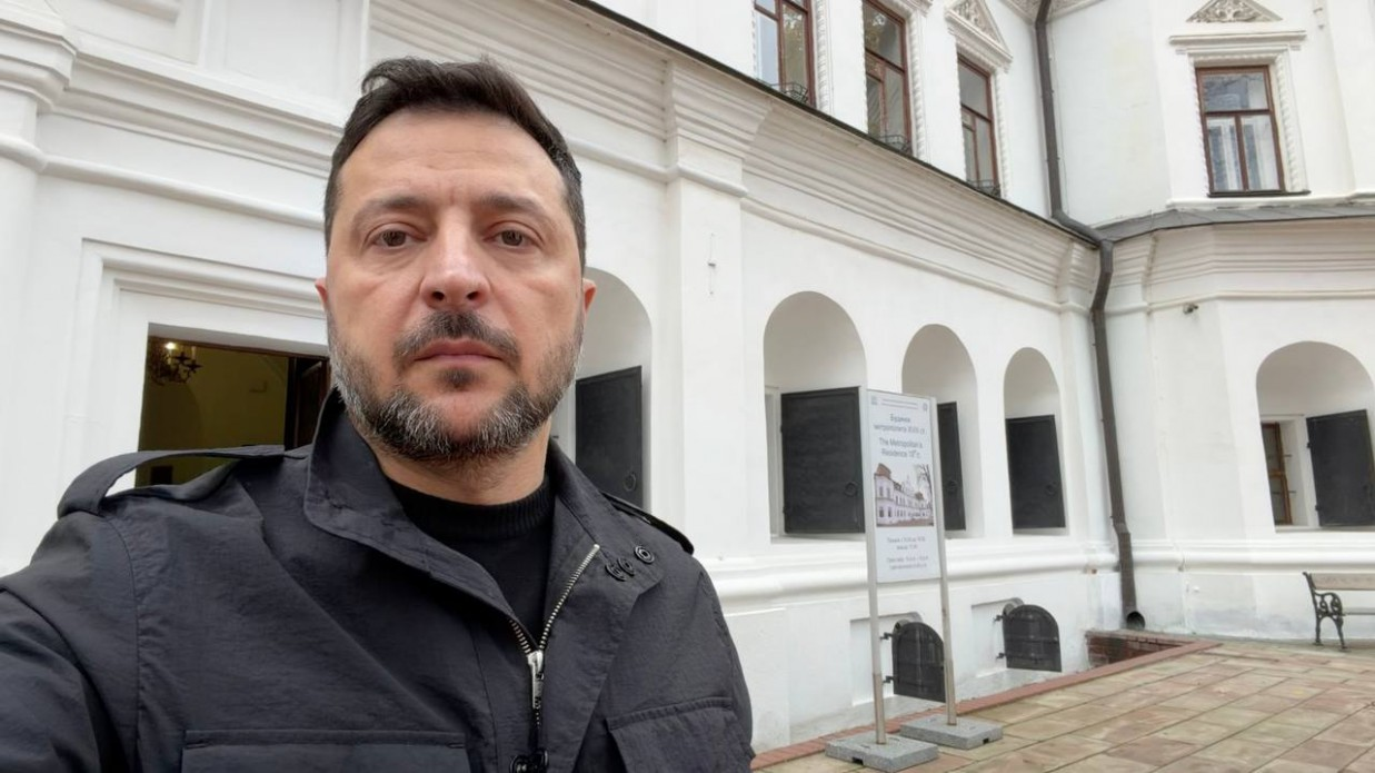 На селфі-світлині крупним планом зображено Президента України Володимира Зеленського під час відеозвернення до українського народу щодо відновлення роботи «Старлінку» та «Старшилду» на колишній швидкості та підготовки гуманітарної операції з розблокування Олешок. Глава держави одягнений у чорну тактичну куртку поверх чорної кофти з високим коміром та має серйозний погляд. На задньому плані праворуч видно велику двоповерхову білу будівлю з арками, коричневими вікнами та зачиненими темними віконницями на першому поверсі.