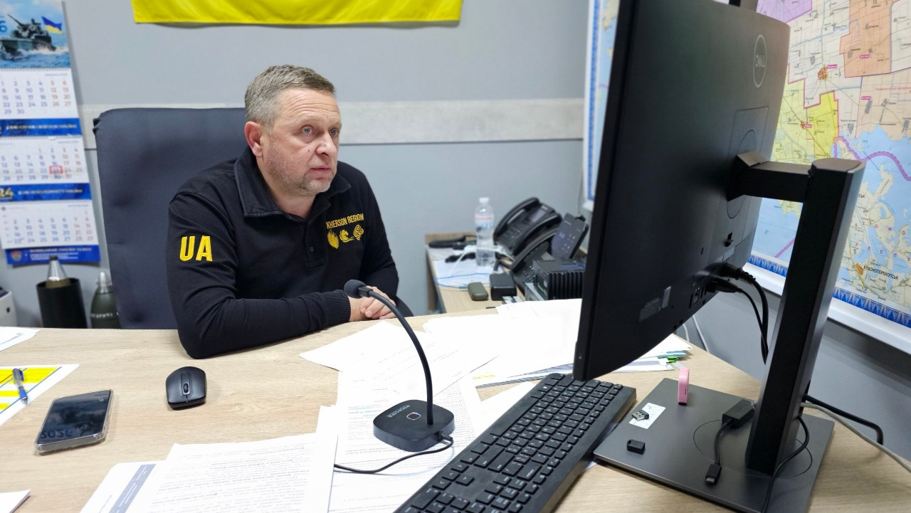 На світлині зафіксовано першого заступника начальника Херсонської ОВА Дмитра Бутрія в робочому кабінеті під час онлайн-народи щодо підготовки до запуску державної програми компенсації аграріям за втрачений через бойові дії врожай. Посадовець сидить у кріслі за кутовим дерев'яним столом у чорній кофті-поло з жовтим написом «UA» на правому рукаві та написом «KHERSON REGION» і гербом області на грудях. Він уважно дивиться на великий чорний монітор комп'ютера, перед яким на столі розкладено робочі документи, бездротову мишу, клавіатуру, настільний мікрофон, флешку та мобільний телефон. На задньому плані кабінету на стіні розвішані велика деталізована карта Херсонської області та державний прапор України, ліворуч висить настінний перекидний календар.