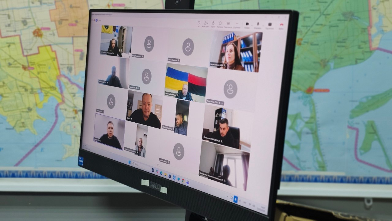 На світлині зафіксовано монітор комп'ютера під час онлайн-наради в Zoom, присвяченої підготовці до запуску державної програми компенсації аграріям за посіви, знищені або не зібрані внаслідок бойових дій. Відеозустріч проходить на тлі великої деталізованої карти Херсонської області, розвішаної на стіні. На екрані відображається сітка з вікнами учасників наради, серед яких керівники військових адміністрацій деокупованих населених пунктів, аграрії та представники профільних установ. У верхньому лівому кутку плитки інтерфейсу чітко видно віконце з кабінету першого заступника начальника ОВА Дмитра Бутрія, а директор Департаменту агророзвитку Дмитро Юнусов підключений в крайньому ліворуч вікні у четвертому ряду знизу, де він сидить у чорній кофті на тлі сірої стіни. 