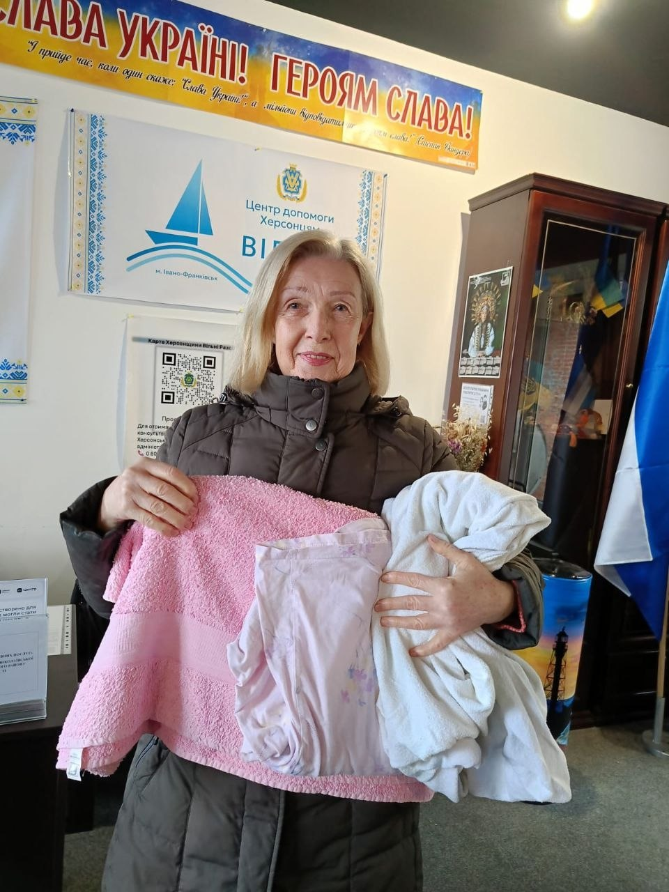 На світлині зафіксовано переселенку з Херсонщини в координаційному центрі «Вільні разом» в Івано-Франківську, яка тримає в руках отриману гуманітарну допомогу, надану за підтримки Фонду громади міста Херсон «Захист». Жінка старшого віку зі світлим волоссям і доброзичливою посмішкою одягнена в темно-коричневу зимову куртку та тримає великий рожевий рушник, світлу постільну білизну й одяг. Вона стоїть у приміщенні хабу на тлі білого банера з синім вітрильником і написом «Центр допомоги Херсонцям ВІЛЬНІ РАЗОМ м. Івано-Франківськ». Угорі на стіні розміщено патріотичний плакат «СЛАВА УКРАЇНІ! ГЕРОЯМ СЛАВА!», праворуч видно темну дерев'яну шафу-вітрину з українським прапором всередині, а ліворуч на стіні наклеєно інформаційний листок із QR-кодом проєкту «Картка Херсонщини Вільні Разом».