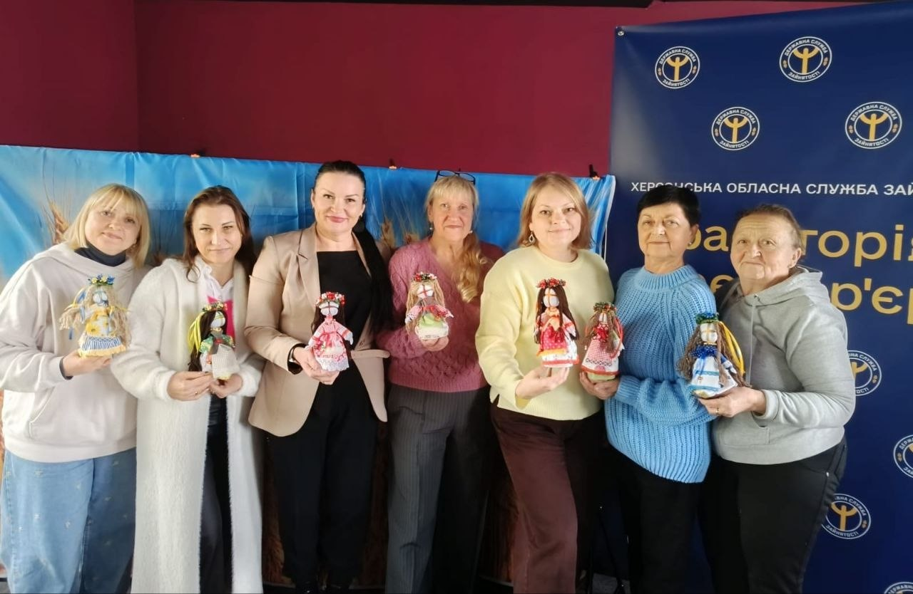 На світлині зображено групу з шести учасниць та майстрині творчого майстер-класу з виготовлення ляльок-мотанок, який відбувся в межах проєкту «Код Херсонщини: пам’ять, культура, єдність» за підтримки Фонду «Захист». Жінки стоять у ряд у світлому приміщенні та з посмішками тримають перед собою власноруч виготовлені традиційні українські обереги в яскравому патріотичному та етнічному вбранні. Учасниці заходу одягнені у повсякденний одяг — світлі худі, довгий білий кардиган, бежевий жакет, рожевий та жовтий светри, а також сіру кофту. Творча зустріч проходить на тлі двох великих банерів: ліворуч розгорнуто блакитно-жовтий стяг із зображенням пшеничного поля, а праворуч — темно-синій інформаційний стенд Херсонської обласної служби зайнятості з написами «Траєкторія твого кар'єрного успіху».