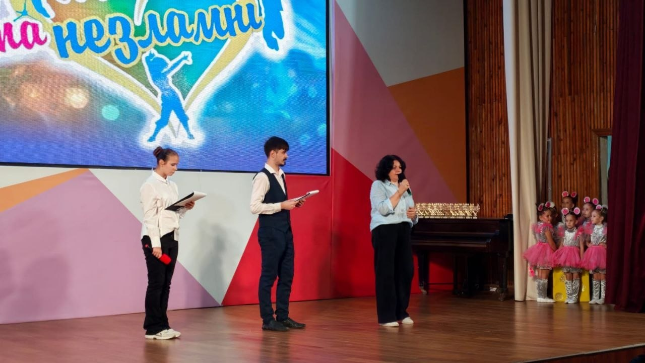 На сцені у Миколаєві під час Міжнародного багатожанрового фестивалю «Талановиті та незламні», організованого Херсонським обласним палацом культури за підтримки Департаменту реалізації гуманітарної політики Херсонської ОДА, стоять троє ведучих: ліворуч — дівчина у білій блузці та чорних штанах з текою і мікрофоном, по центру — чоловік у білій сорочці та чорній жилетці з папкою в руках, праворуч — директорка Департаменту реалізації гуманітарної політики Херсонської ОДА Віолета Царькова у блакитній сорочці, яка виступає перед публікою. Ззаду на великому світлодіодному екрані відображається яскравий банер проєкту з написом «Талановиті та незламні» та силуетом дівчини, що танцює. У правій частині сцени, біля піаніно, виступу очікує група маленьких учасниць у рожевих танцювальних костюмах, які представляють творчість та культурні традиції незламного півдня України.