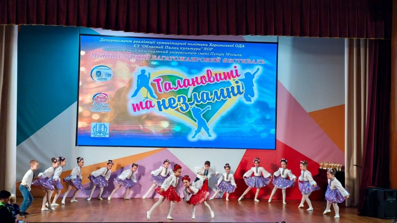 На сцені у Миколаєві під час Міжнародного багатожанрового фестивалю «Талановиті та незламні», організованого Херсонським обласним палацом культури за підтримки Департаменту реалізації гуманітарної політики Херсонської ОДА, виступає дитячий танцювальний колектив. Юні артисти виконують динамічну народну хореографічну постановку: дівчатка одягнені у вишиванки та блискучі пишні спідниці (більшість у фіолетових, а дві солістки по центру — в яскраво-червоних), хлопчики — у білих сорочках та темних штанах. Ззаду на великому світлодіодному екрані відображається яскравий банер проєкту з логотипами організаторів, написом «Міжнародний багатожанровий фестиваль "Талановиті та незламні"» та кольоровими силуетами дітей, що танцюють. Цей виступ є частиною масштабної програми, яка об'єднала понад 300 виконавців різного віку задля збереження українських культурних традицій та представлення творчої спадщини півдня країни.