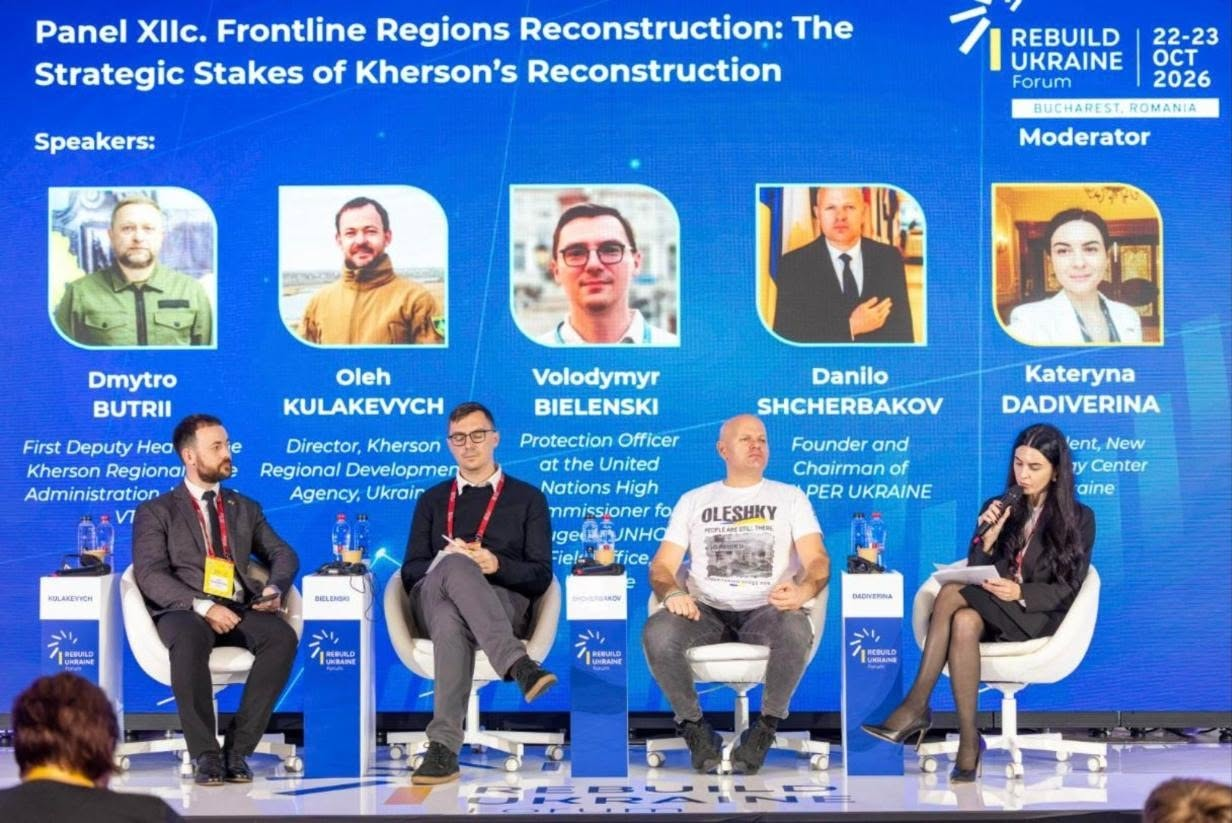 На сцені міжнародного форуму «Rebuilding Ukraine» у Бухаресті під час панельної дискусії «Frontline Regions Reconstruction: The Strategic Stakes of Kherson’s Reconstruction» на білих кріслах сидять спікери та модераторка, а перед кожним із них встановлено невелику білу інформаційну тумбу з мікрофоном. Зліва направо на сцені перебувають: директор Агенції регіонального розвитку Херсонщини Олег Кулакевич у чорному костюмі та білій сорочці з жовтим бейджем, офіцер із питань захисту УВКБ ООН Володимир Бєленський у сірому костюмі, окулярах та червоній краватці, засновник організації «PRO UKRAINE» Данило Щербаков у сірих джинсах та білій футболці з принтом «OLESHKY», а також представниця New Strategy Center Катерина Дадіверіна в чорному вбранні, яка тримає в руках папери. Ззаду на великому яскраво-синьому екрані відображаються офіційні логотипи заходу з датами «22-23 OCT 2026», назва дискусійної панелі, позначки «Speakers» та «Moderator», а також п'ять портретних фотографій імена учасників, серед яких першим зазначено першого заступника начальника Херсонської ОВА Дмитра Бутрія.