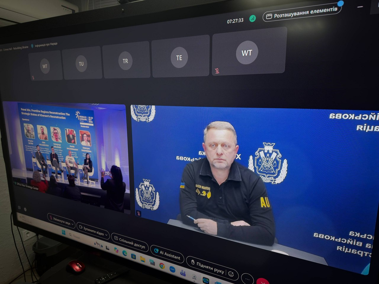 На екрані комп'ютера відображається інтерфейс онлайн-конференції, під час якої перший заступник начальника Херсонської ОВА Дмитро Бутрій бере дистанційну участь у міжнародному форумі «Rebuilding Ukraine» у Бухаресті. Права, більша частина вікна трансляції показує Дмитра Бутрія, який сидить у чорній форменій сорочці з нашивкою «Херсонська ОВА» на тлі брендованого синього банера з офіційними гербами Херсонської області. У лівій частині екрана у меншому вікні виведено пряме включення із зали засідань у Румунії, де триває панельна дискусія «Frontline Regions Reconstruction: The Strategic Stakes of Kherson’s Reconstruction» за участі інших спікерів, а на задньому плані сцени видно великий синій банер форуму. У верхній та нижній частинах монітора розташовані елементи керування онлайн-платформою для зв'язку, зокрема кнопки «Спільний доступ», «AI Assistant» та «Підняти руку».