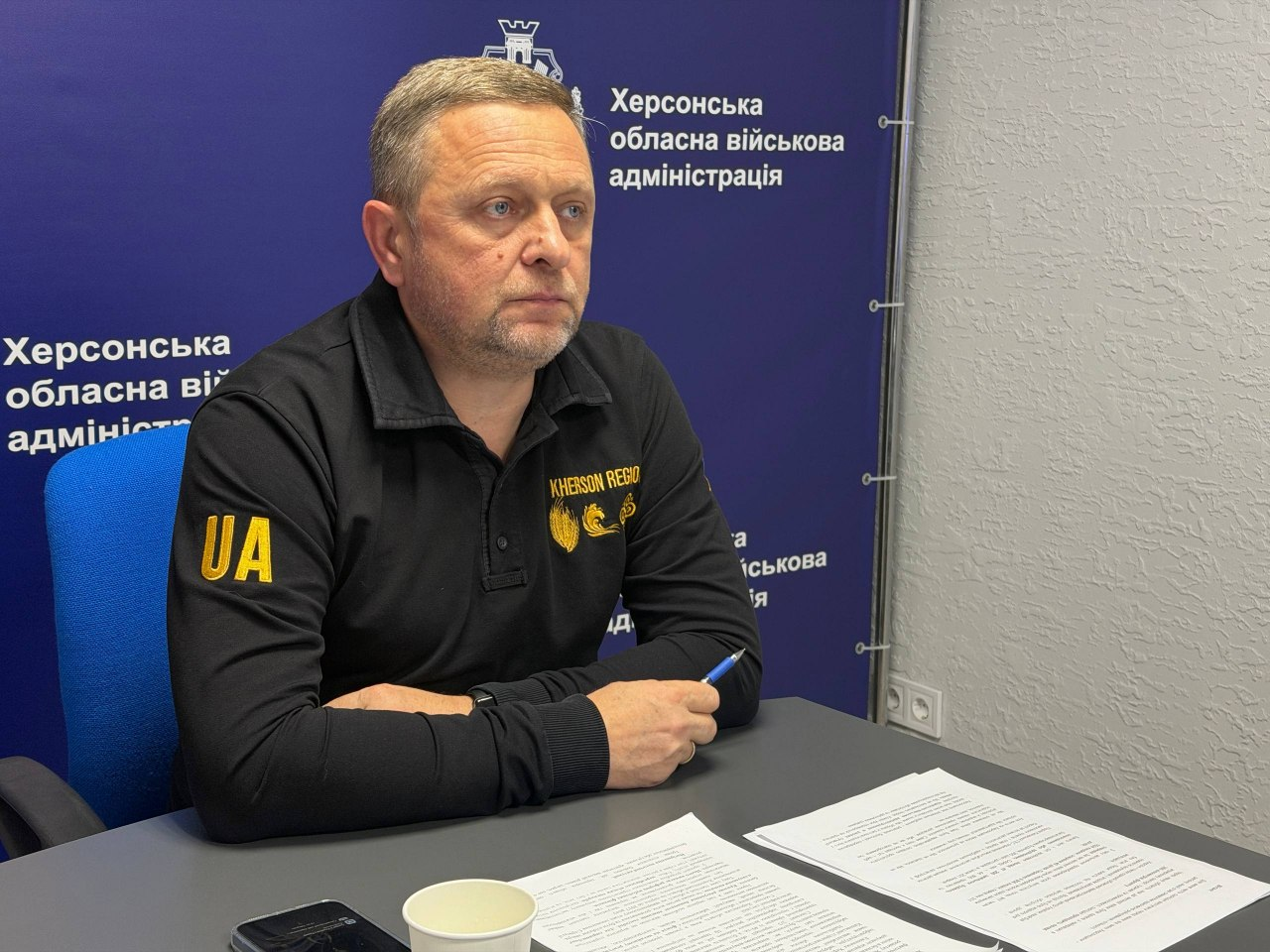 На фоні брендованого темно-синього банера з офіційним гербом та написом «Херсонська обласна військова адміністрація» за столом сидить перший заступник начальника Херсонської ОВА Дмитро Бутрій. Він одягнений у чорну кофту із жовтою абревіатурою «UA» на правому плечі та вишитим золотистим гербом області й написом «KHERSON REGION» на грудях. Посадовець сидить у півоберта, тримаючи в правій руці ручку, а перед ним на столі лежать роздруковані документи з текстом оновленої Стратегії розвитку Херсонщини, паперовий стаканчик та мобільний телефон. Цей кадр зафіксував процес його дистанційної участі та онлайн-виступу на міжнародному форумі «Rebuilding Ukraine» у Бухаресті, де обговорювалися стратегічні підходи до відновлення прифронтових територій півдня України.