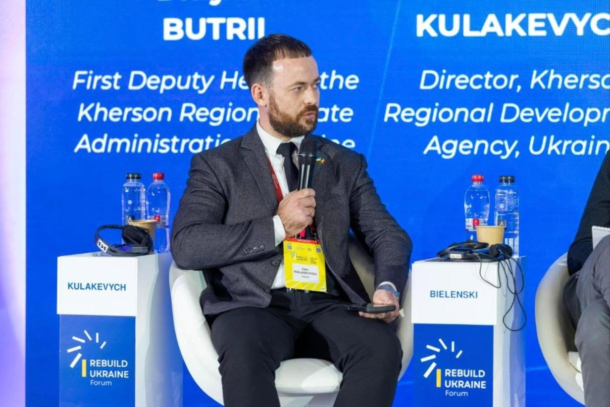 На сцені міжнародного форуму «Rebuilding Ukraine» у Бухаресті під час панельної дискусії, присвяченої відбудові прифронтових регіонів, у білому кріслі сидить директор Агенції регіонального розвитку Херсонщини Олег Кулакевич. Він одягнений у класичний темно-сірий піджак, білу сорочку та темну краватку, а на його піджаку закріплено яскраво-жовтий інформаційний бейдж учасника заходу. Посадовець тримає в руці мікрофон і виступає перед присутніми, повернувши голову ліворуч. Обабіч його крісла встановлено дві невеликі білі брендовані тумби з логотипом форуму «Rebuilding Ukraine Forum» та написами «KULAKEVYCH» і «BIELENSKI», на яких стоять пляшки з водою та навушники для синхронного перекладу. Ззаду на великому яскраво-синьому світлодіодному екрані відображаються великі англомовні написи з іменами спікерів та їхніми посадами, зокрема фрагменти тексту «BUTRII First Deputy Head of the Kherson Region Administration» та «KULAKEVYCH Director, Kherson Regional Development Agency, Ukraine».