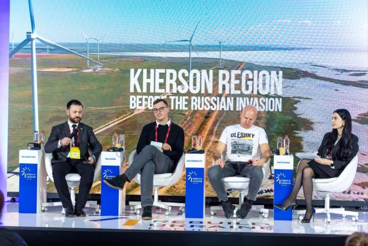 На сцені міжнародного форуму «Rebuilding Ukraine» у Бухаресті під час панельної дискусії «Frontline Regions Reconstruction: The Strategic Stakes of Kherson’s Reconstruction» на чотирьох білих кріслах сидять спікери та модераторка, а перед кожним із них встановлено невелику білу інформаційну тумбу з мікрофоном. Зліва направо на сцені перебувають: директор Агенції регіонального розвитку Херсонщини Олег Кулакевич у чорному костюмі та білій сорочці з жовтим бейджем, офіцер із питань захисту УВКБ ООН Володимир Бєленський у чорному джемпері, окулярах та червоній стрічці-бейджу, засновник організації «PRO UKRAINE» Данило Щербаков у сірих джинсах та білій футболці з принтом «OLESHKY», а також представниця New Strategy Center Катерина Дадіверіна в чорній сукні та туфлях-човниках, яка тримає в руках папери. Ззаду на великому світлодіодному екрані відображається панорамна світлина вітрової електростанції на узбережжі Херсонщини з великим білим англомовним написом «KHERSON REGION BEFORE THE RUSSIAN INVASION». На передньому плані перед кріслами видно край сцени з частиною дзеркального логотипа форуму.