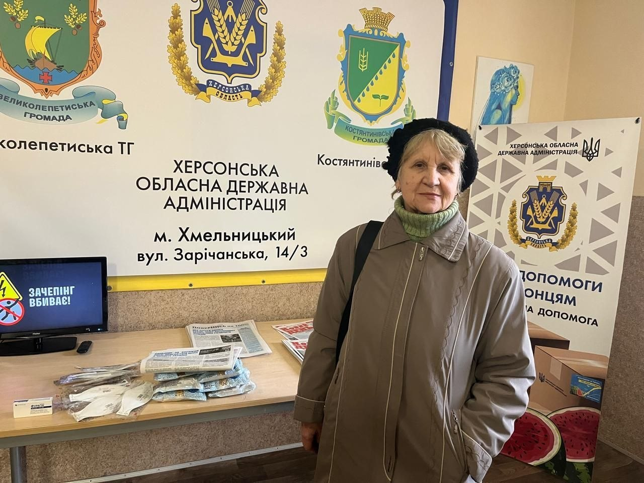 У приміщенні координаційного центру «Вільні разом» у Хмельницькому на тлі інформаційних стендів стоїть літня жінка у світло-коричневій куртці, зеленому светрі з високим коміром та чорному береті, яка дивиться в кадр. Ззаду неї на стіні розміщено великий білий банер із гербами Великолепетиської та Костянтинівської громад, офіційним гербом Херсонської області та написом «Херсонська обласна державна адміністрація, м. Хмельницький». Праворуч встановлено вертикальний банер «Центр допомоги херсонцям», а ліворуч стоїть стіл, на якому розкладено примірники свіжих друкованих газет «Новий день» та інша друкована продукція для переселенців. Також ліворуч на столі видно край увімкненого монітора комп'ютера із застережливим патріотичним заставним екраном «Зачепінг вбиває!».