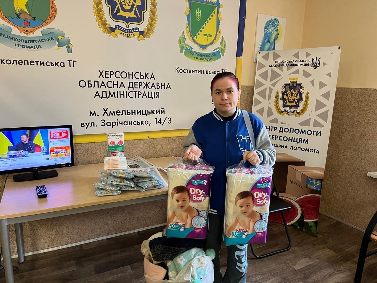 У приміщенні координаційного центру «Вільні разом» у Хмельницькому молода жінка-переселенка з Херсонщини в синій спортивній кофті з білими смугами на рукавах та сірих спортивних штанях стоїть перед великим інформаційним банером, тримаючи в руках дві великі пачки дитячих підгузків бренду «Dry & Soft». Ззаду неї на стіні розміщено банер із гербами Великолепетиської та Костянтинівської громад, офіційним гербом Херсонської області та написом «Херсонська обласна державна адміністрація, м. Хмельницький». Праворуч встановлено вертикальний банер «Центр допомоги херсонцям. Гуманітарна допомога», а ліворуч стоїть стіл, на якому розкладено свіжу пресу. Також ліворуч на столі видно монітор комп'ютера, на екрані якого транслюється телевізійний сюжет інформаційного марафону.