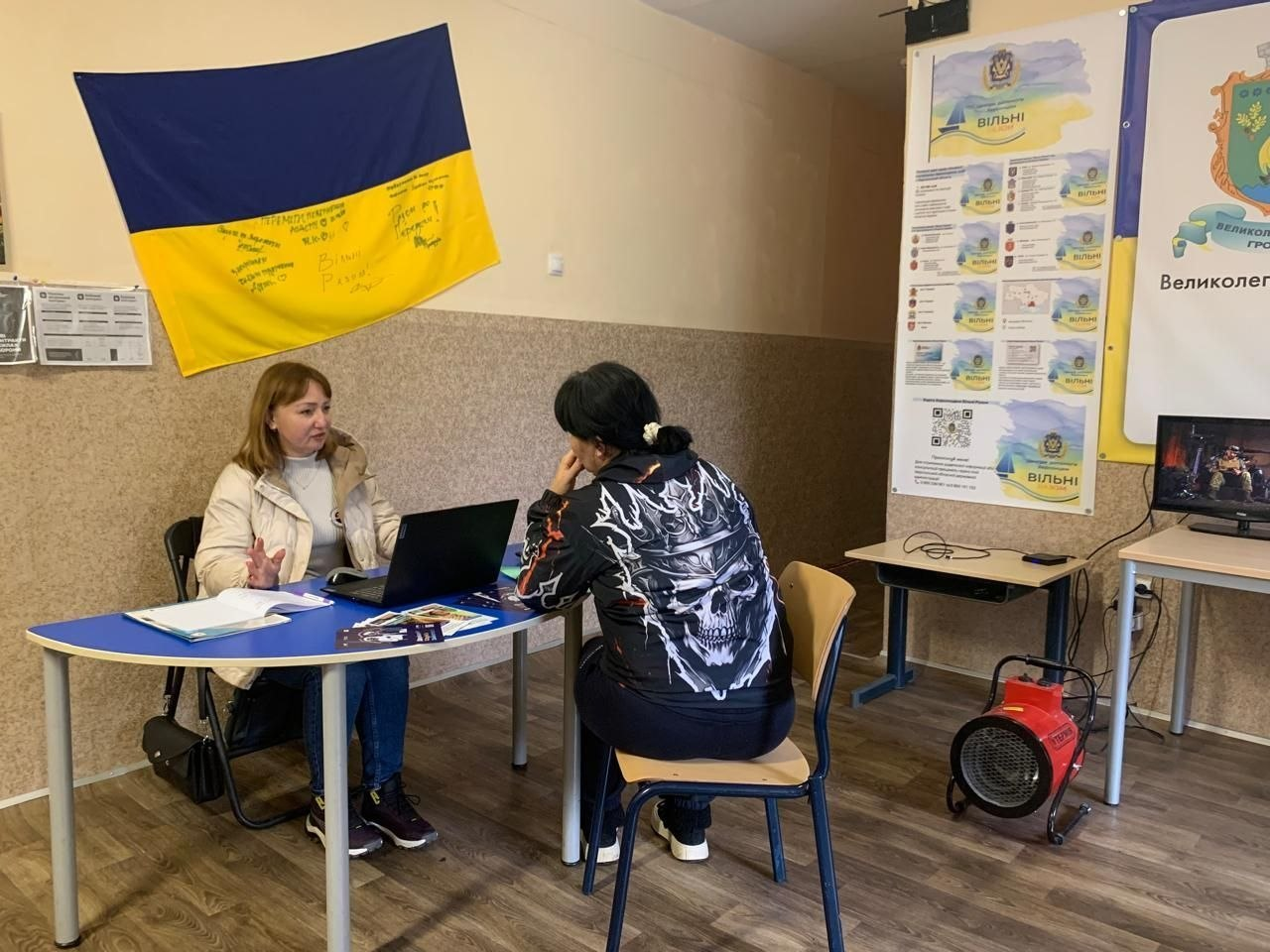 У приміщенні координаційного центру «Вільні разом» у Хмельницькому за синім напівкруглим столом сидить фахівчиня центру зайнятості у світлій куртці та синіх джинсах, яка надає консультацію з питань працевлаштування відвідувачці — жінці з темним волоссям у чорній кофті з великим білим принтом у вигляді черепа в короні на спині. На столі перед ними відкритий чорний ноутбук та розкладені інформаційні буклети. Ліворуч на стіні кабінету висить державний прапор України з патріотичними підписами та побажаннями від захисників, а праворуч на стендах розміщено інформаційні плакати проєкту «Вільні» та частину банера з гербом Великолепетиської громади. Поруч на столі видно увімкнений монітор комп'ютера, а на підлозі біля стільця стоїть червоний промисловий обігрівач-теплова гармата.