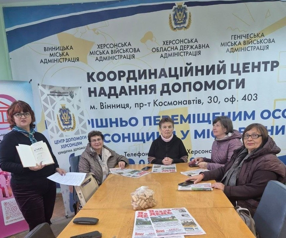 У приміщенні Вінницького координаційного центру під час літературної години, присвяченої Дню української писемності та мови, на тлі великого офіційного банера з гербом області та написом «КООРДИНАЦІЙНИЙ ЦЕНТР НАДАННЯ ДОПОМОГИ» за довгим дерев'яним столом сидить група жінок-переселенок з Херсонщини. Ліворуч стоїть провідниця заходу в окулярах та чорній кофті з синьою шийною хусткою, яка демонструє присутнім розгорнуту білу книгу. За столом сидить чотири жінки у повсякденному зимовому одязі та куртках, які уважно слухають лекторку. На столі перед ними розкладено свіжі примірники газет «Новий день» та лежить прозорий пакет із частуваннями.