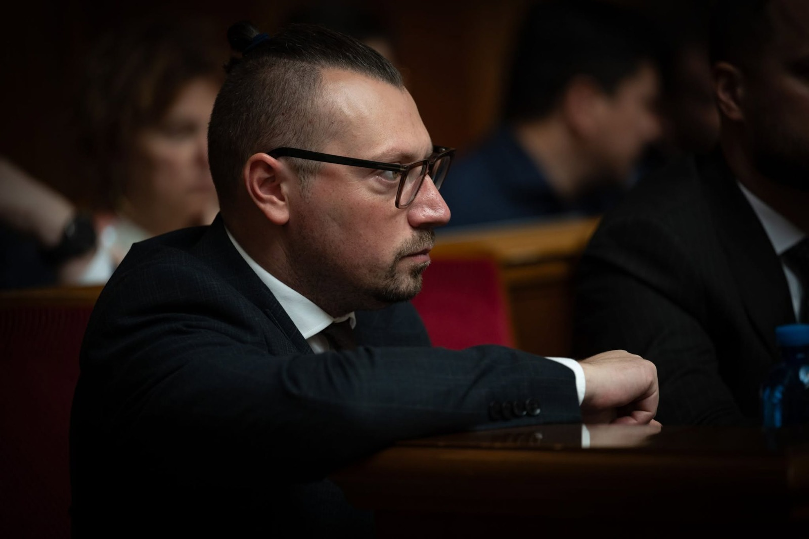 У залі засідань Верховної Ради України великим планом у профіль зафіксовано Міністра у справах громад, територій та внутрішньо переміщених осіб України Віталія Безгіна, який сидить на своєму місці. Він одягнений у класичний чорний костюм, білу сорочку та краватку, носить окуляри в темній оправі. Посадовець зосереджено поклав руку на край дерев'яного столу сесійної зали й уважно слухає виступи. На задньому плані в розмитті помітно силуети інших учасників засідання та дерев'яне оздоблення стін.
